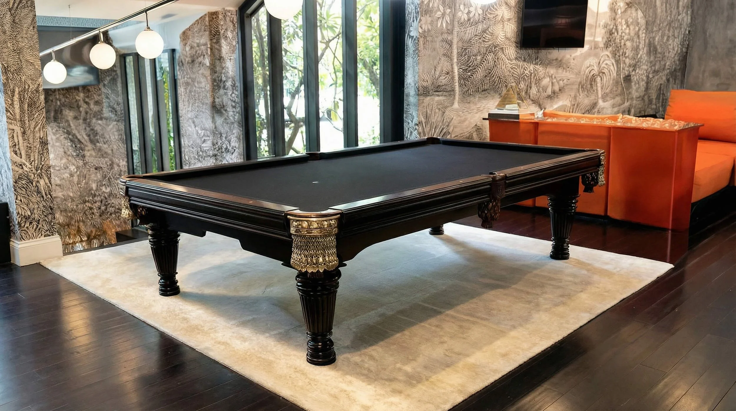 A black pool table with ornate legs sits on a light-colored rug in a room with dark wood flooring. Behind it, there are large windows, bushes outside, and a patterned wall with foliage designs. To the right, there is an orange couch and a small table