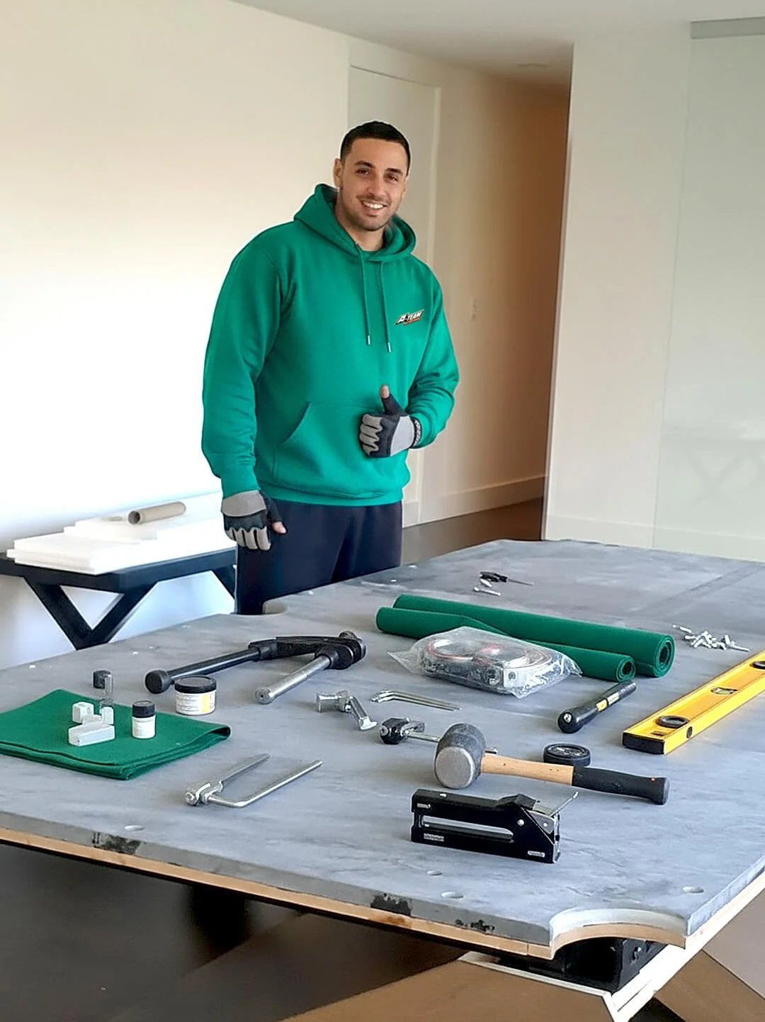 A man smiling at a work table with various tools, rolls of green material, gloves, and other equipment. He is wearing a teal hoodie and black gloves, standing indoors in a room with a white wall.