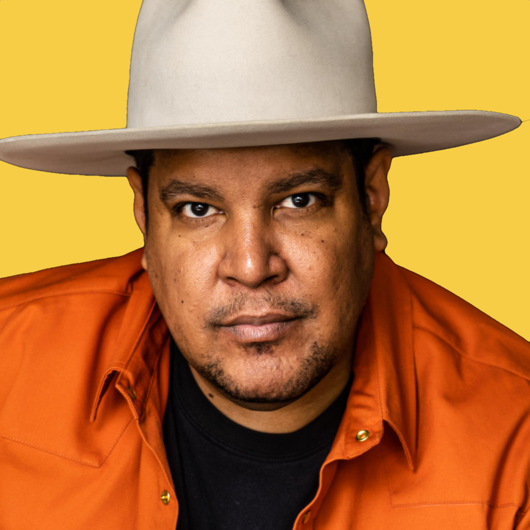 A man wearing a wide-brimmed beige hat, an orange shirt, and a black shirt underneath, looking directly at the camera against a yellow background.