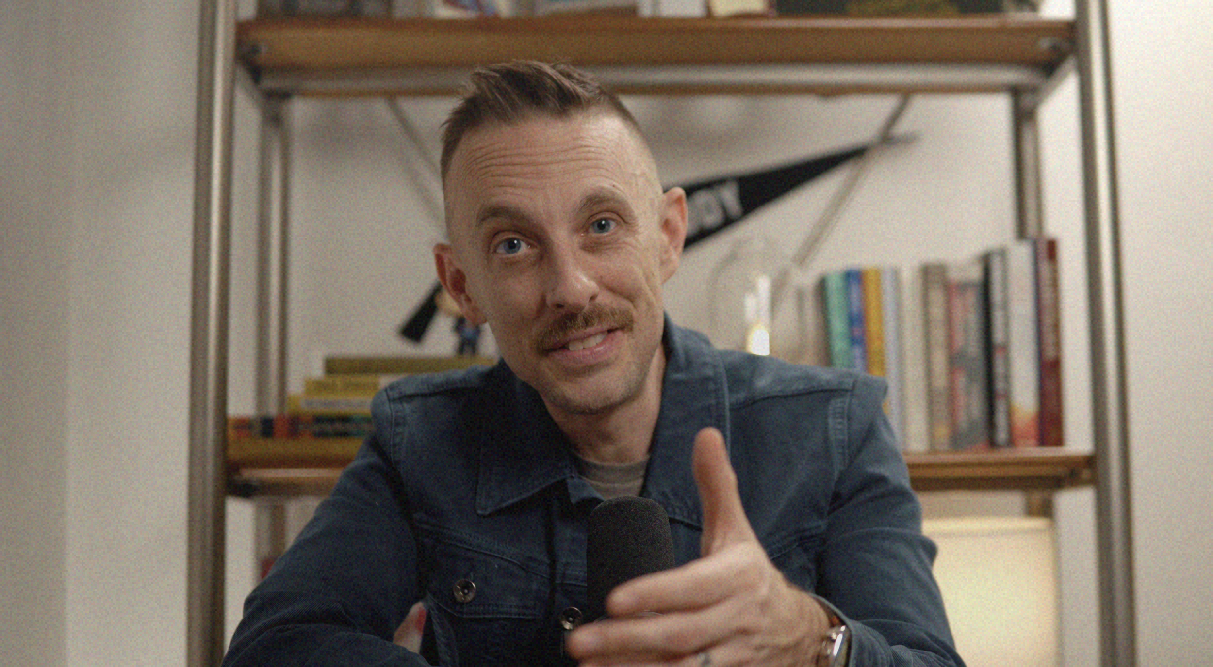 A man with short hair, a mustache, and a goatee, wearing a blue jacket, is holding a microphone and making a thumbs-up gesture. Behind him is a bookshelf with books and a white wall.
