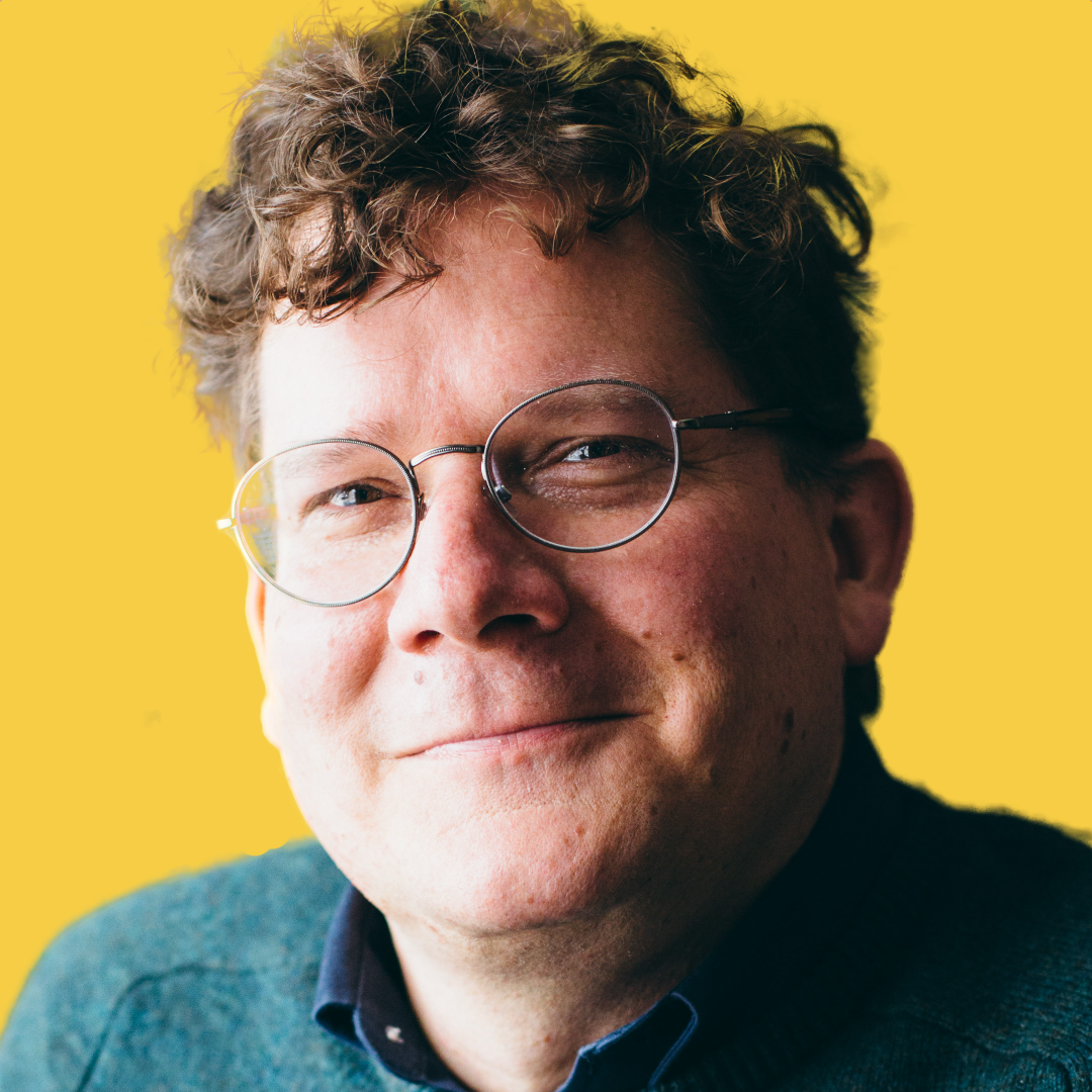 Close-up of a smiling man with curly hair and glasses against a yellow background.