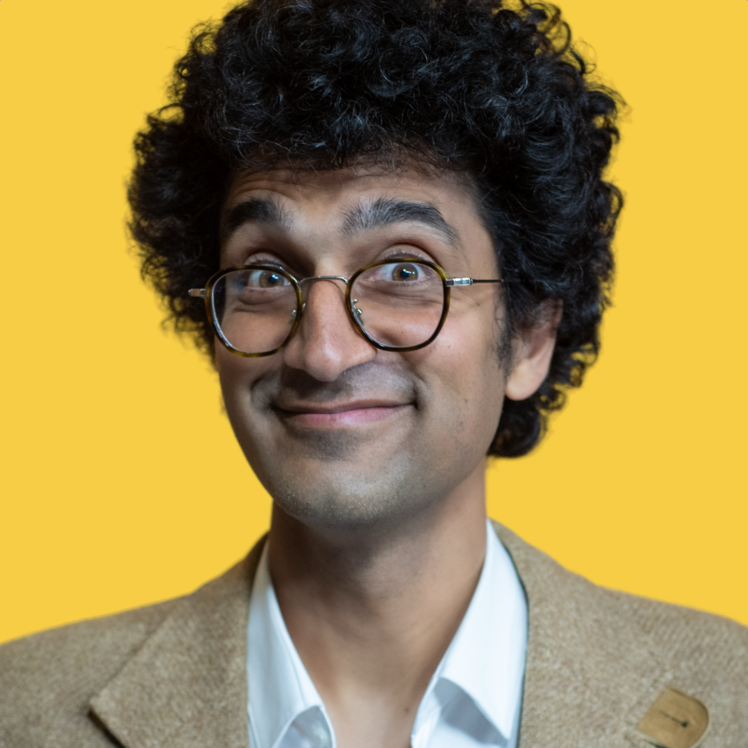 Smiling man with glasses and curly hair wearing a suit jacket against a yellow background.