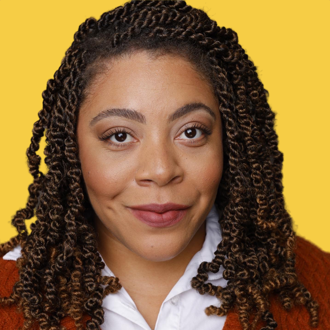 Close-up portrait of a woman with dark brown twisted hair and light makeup, wearing a white shirt and a brown sweater against a yellow background.