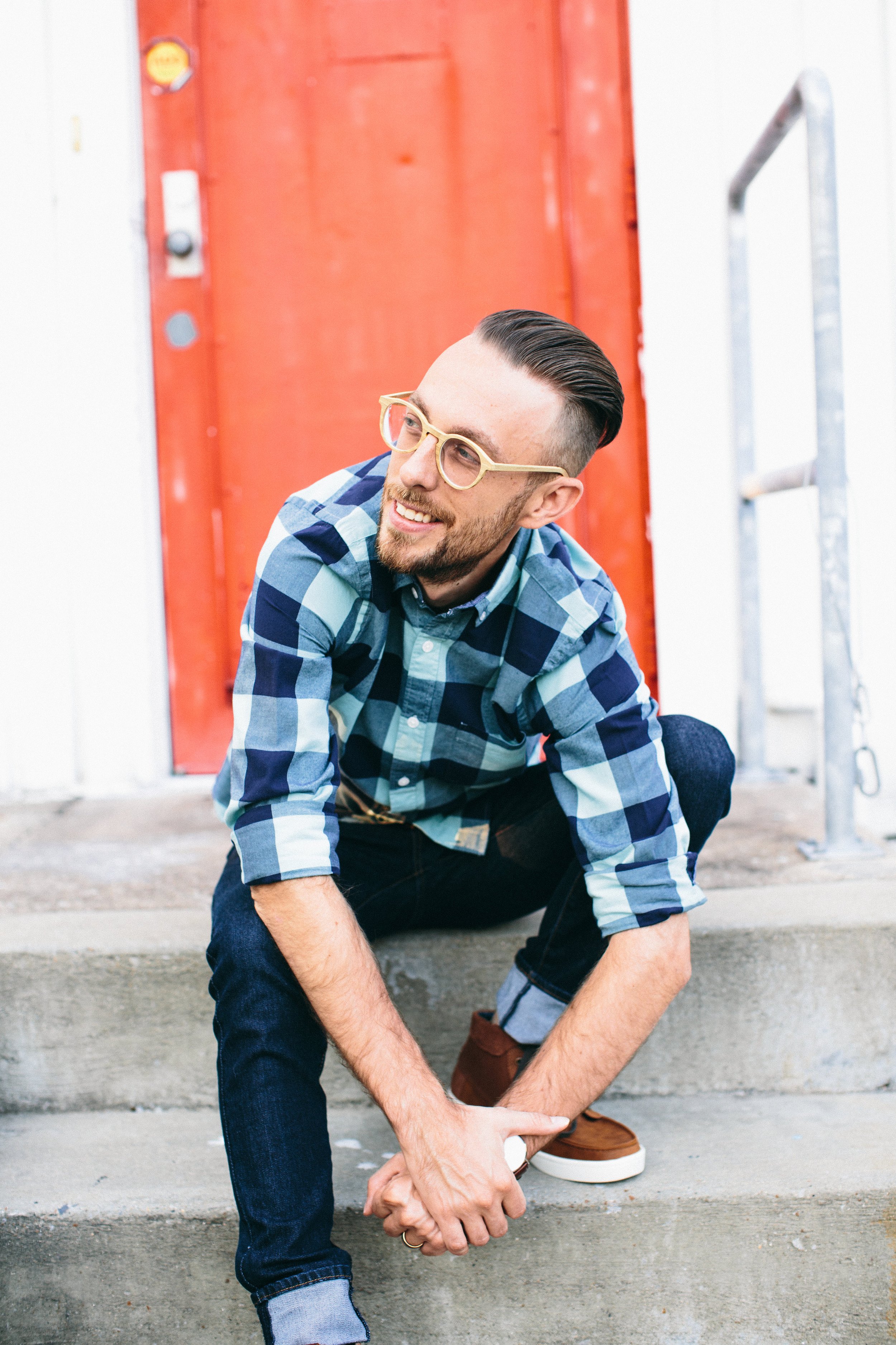 A man with glasses and a beard crouching on a concrete step, tying his shoelace in front of a red door.