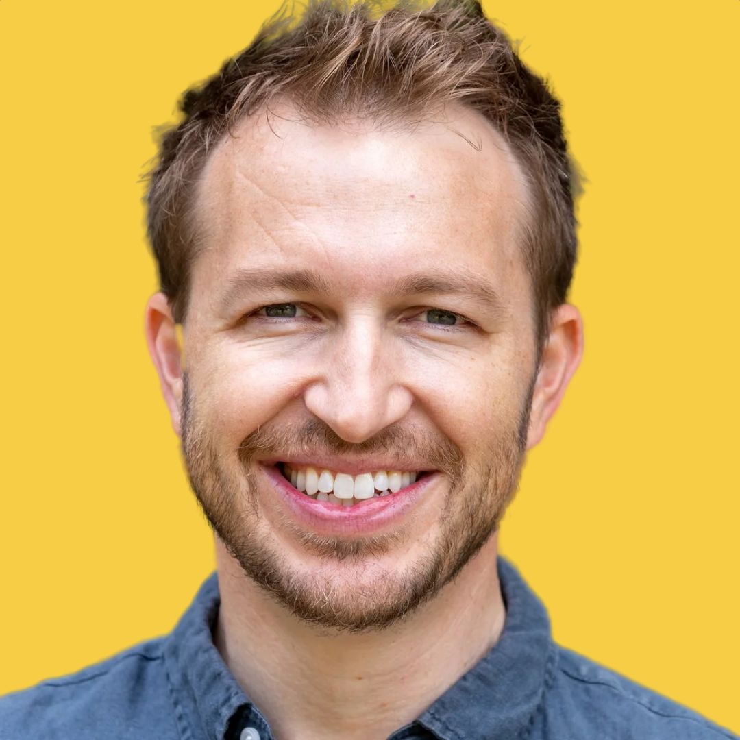 A smiling man with short brown hair, a beard, and blue eyes, wearing a blue shirt against a yellow background.