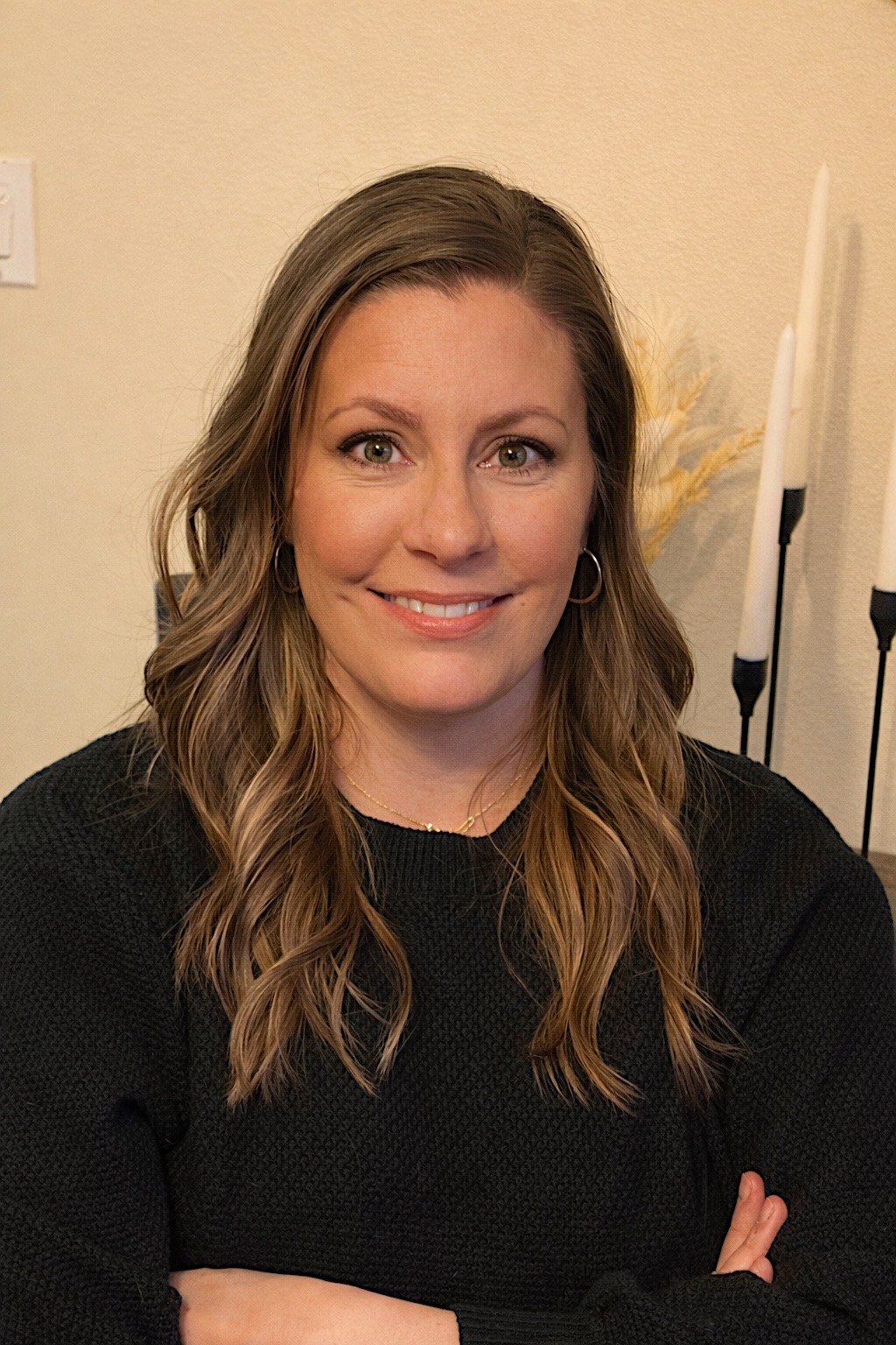 A woman with wavy, shoulder-length brown hair, green eyes, and light skin, smiling with crossed arms, wearing a black sweater and small hoop earrings, indoors with beige walls and white candles in the background.