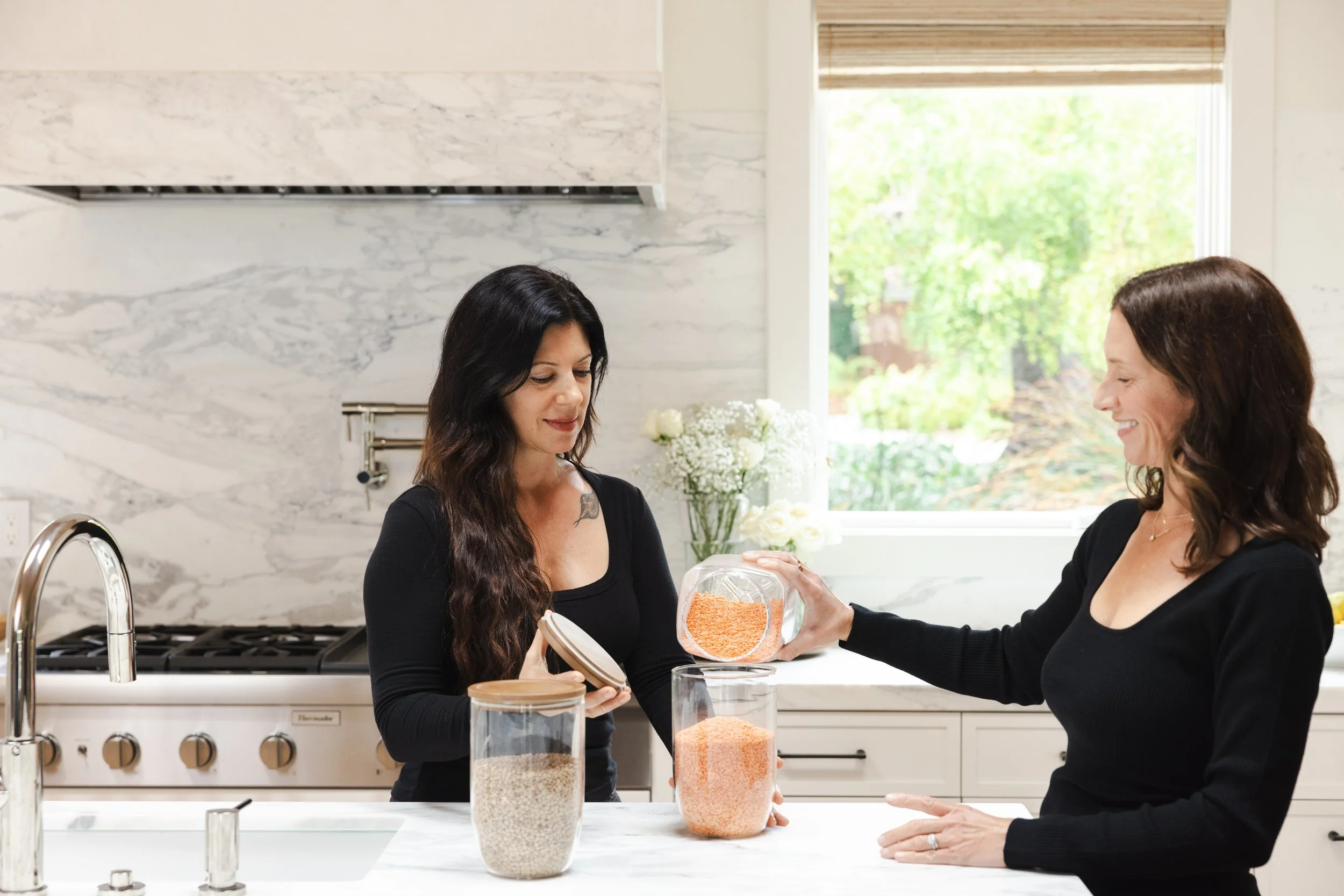 Two women organizing pantry staples in a bright modern kitchen while exchanging a glass jar of orange lentils.