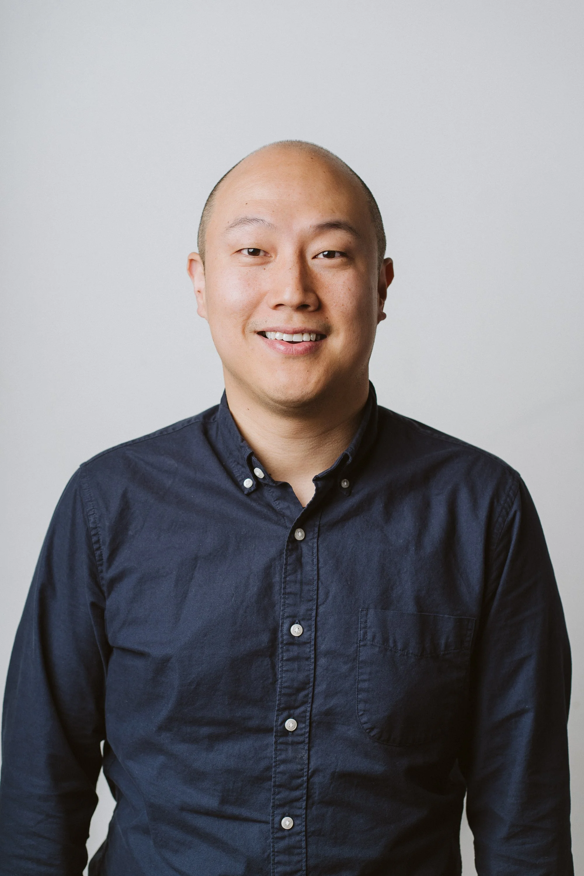 A young man with a bald head and light skin, smiling, wearing a dark button-up shirt, against a plain light gray background.