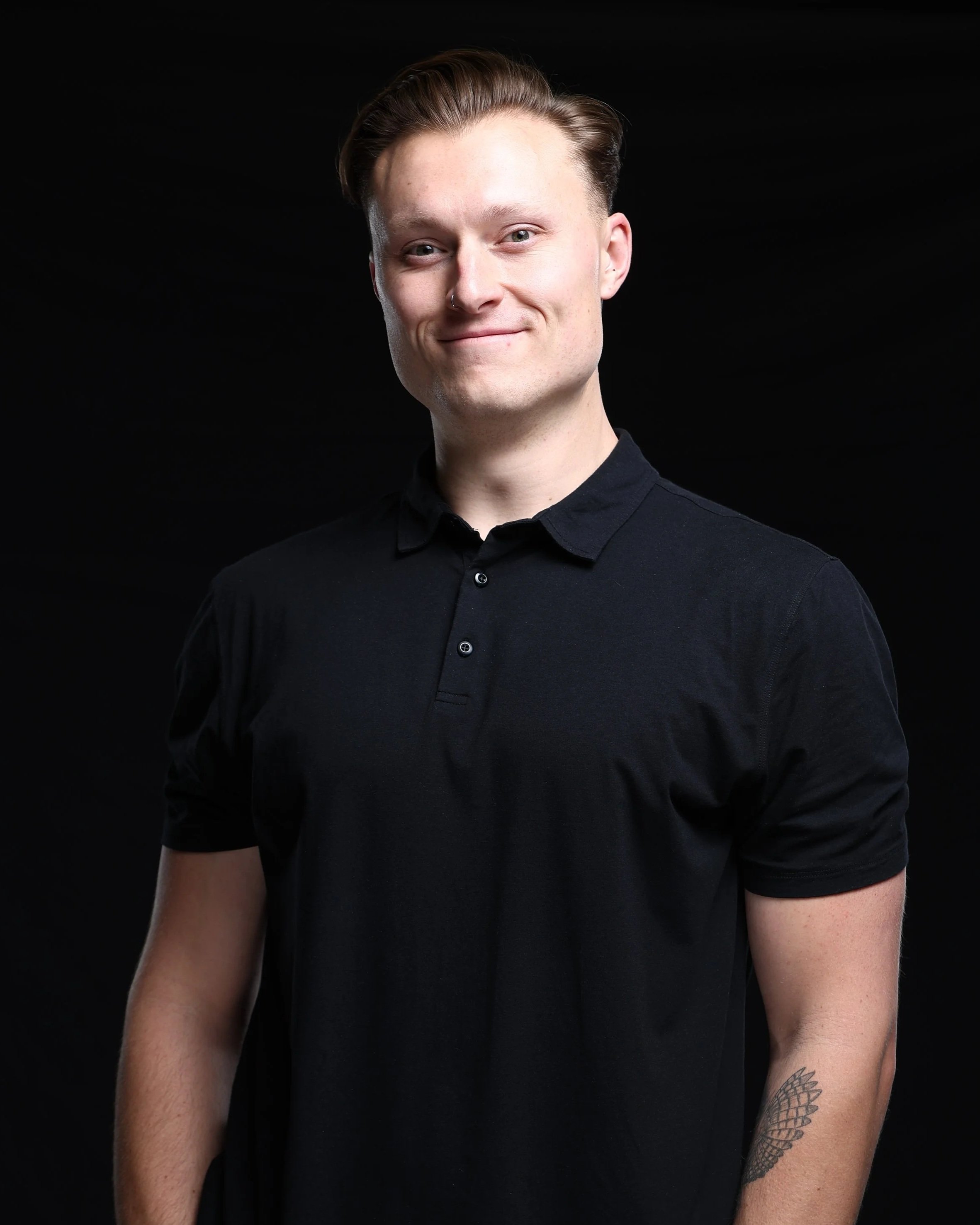 A young man with short, light brown hair wearing a black polo shirt, smiling, standing against a black background.