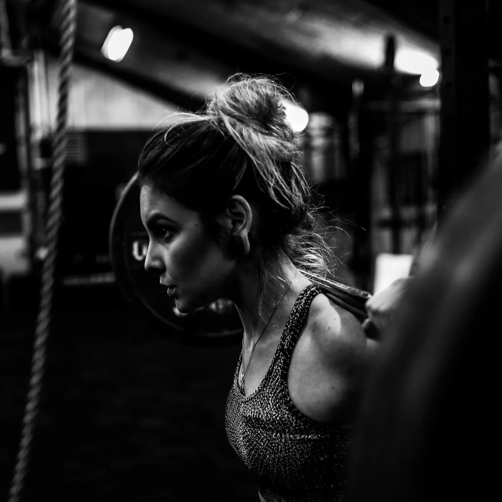 A woman with her hair in a messy bun, wearing a tank top, appearing focused during a workout in a gym with weights in the background.
