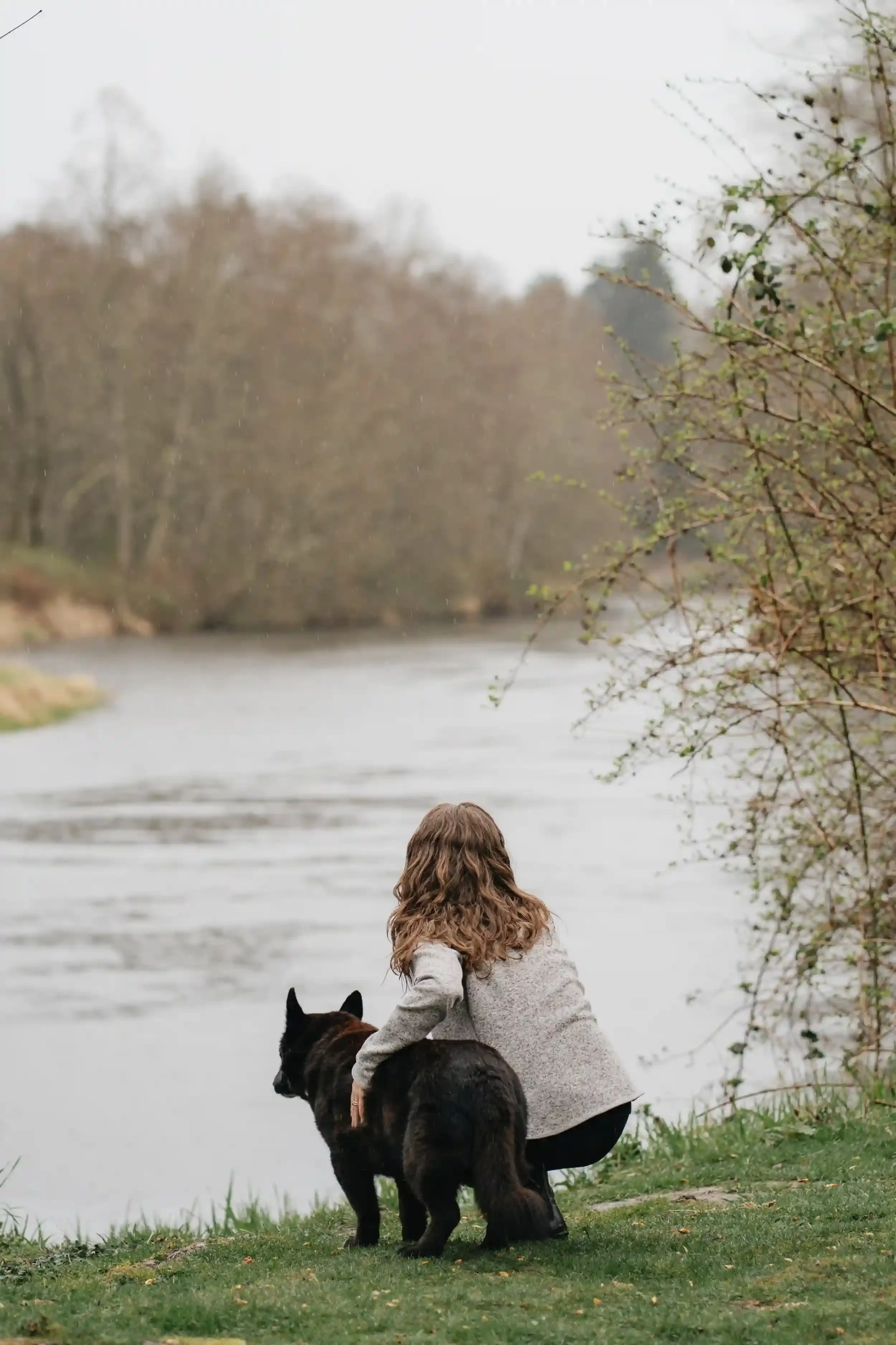 A woman with long, wavey hair is squatting near a short black dog by a river, with trees and overcast sky in the background.