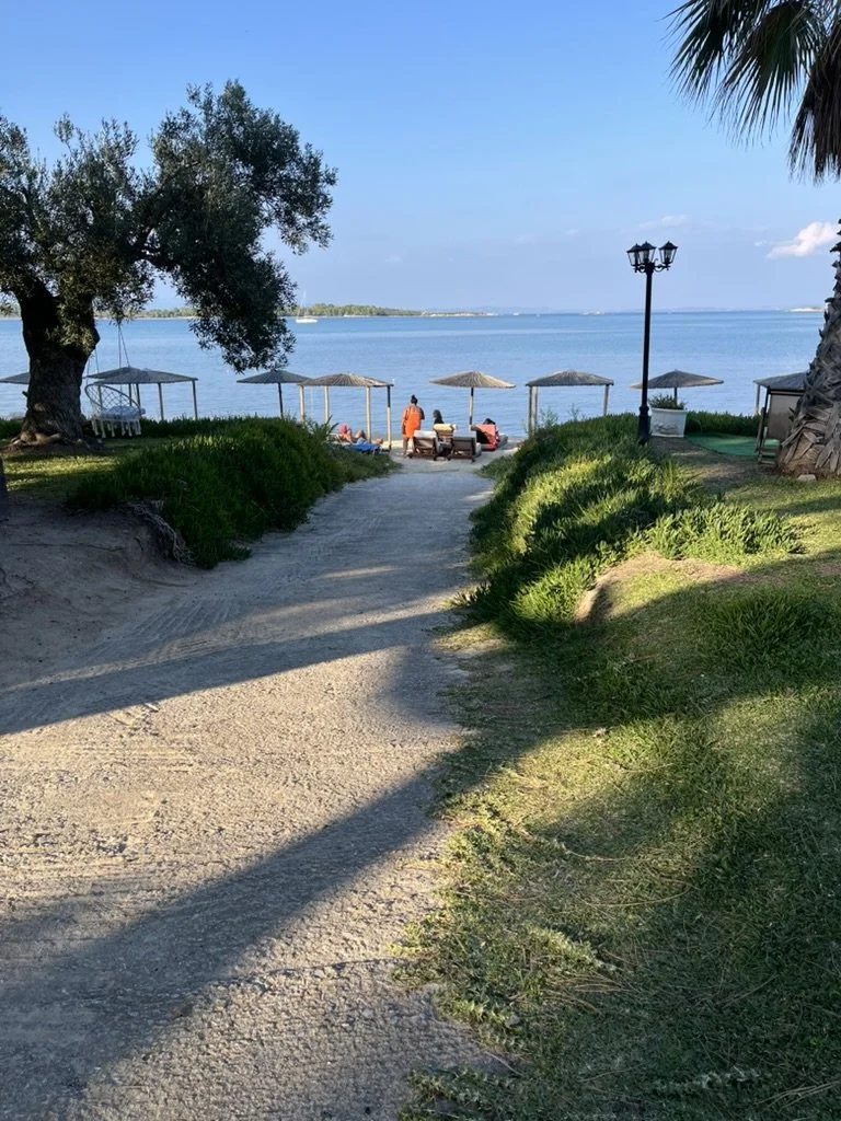 A sandy pathway leading to a beach area with umbrellas, lounge chairs, and people enjoying the water. There are trees and greenery on either side, and the ocean is visible in the background under a clear blue sky.