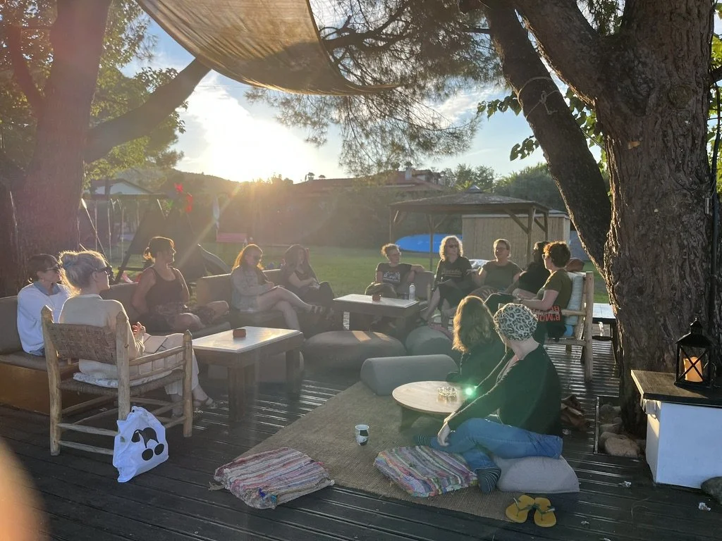 Writers' workshop in Sithonia, Halkidiki, Greece with Ink and Salt. Writers sitting outdoors on wooden deck under large trees in the sunlight, having a gathering or discussion.