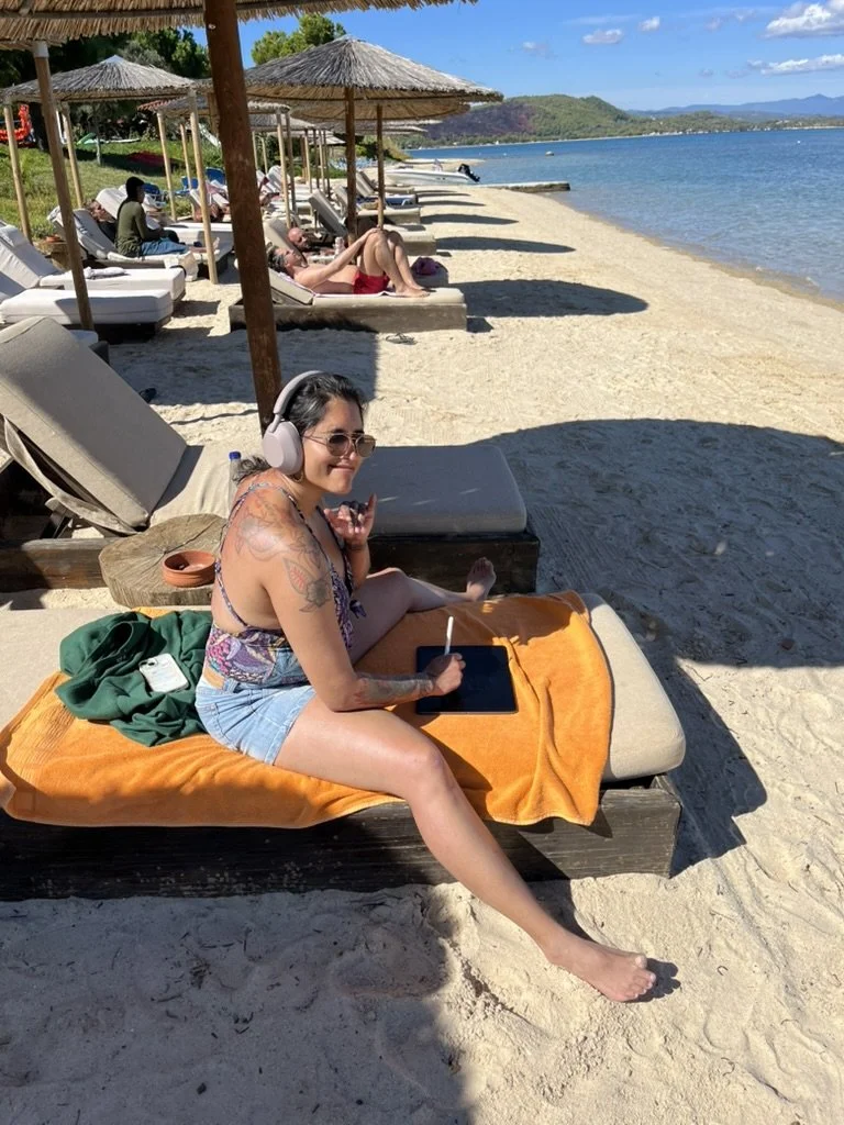 Writer relaxing on a lounge chair at a beach, wearing headphones and sunglasses, with a tablet and stylus, other beachgoers under umbrellas, sandy shore, calm sea, mountains in the background. Ink and Salt writing retreats