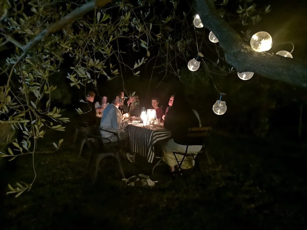 Group of writers reading from their work sitting around a table outdoors at night, illuminated by string lights and lanterns, surrounded by trees.
