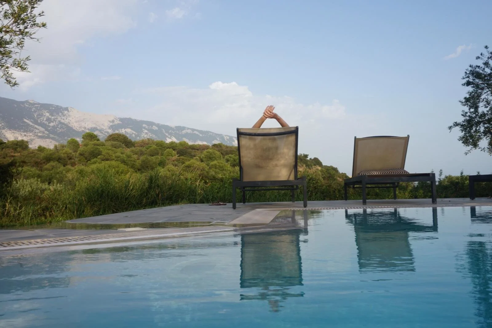 A swimming pool with two lounge chairs overlooking a lush green landscape and mountains in the background under a partly cloudy sky at an Ink and Salt writing retreat