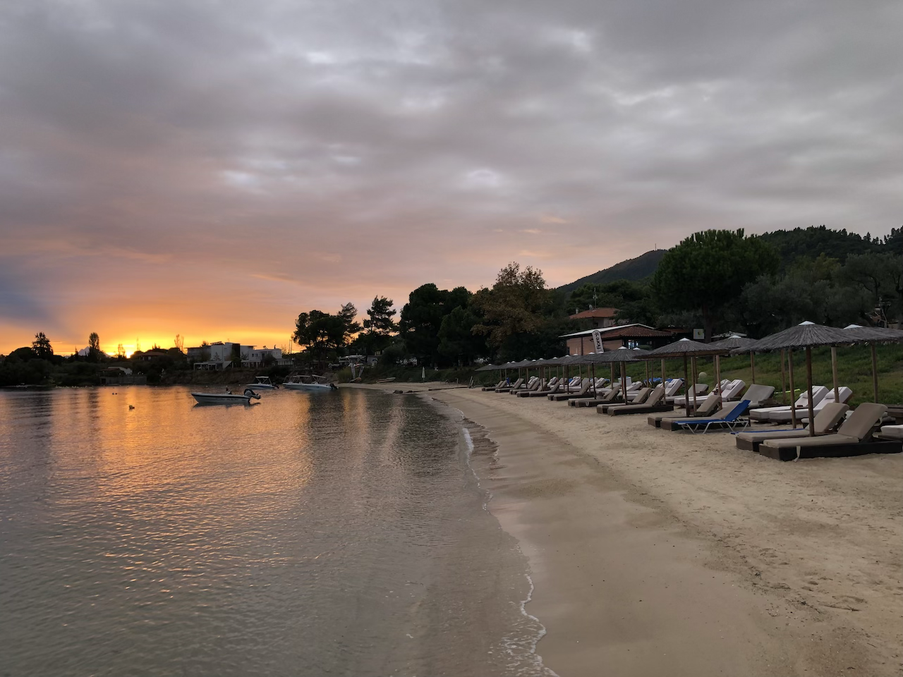 A serene lakeside beach at sunset with empty lounge chairs and umbrellas along the sandy shore, calm water reflecting the sky, and a distant mountainous landscape under a cloudy sky.