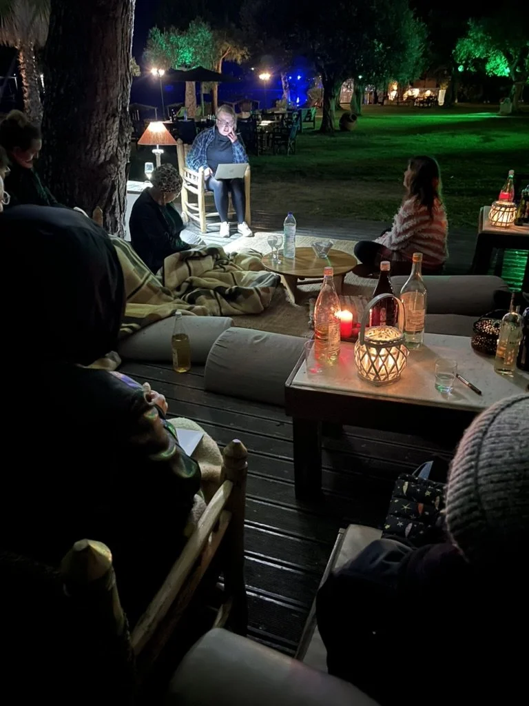 People sitting outside at night around a table with drinks, blankets, and candles, with trees and string lights in the background, while one person is using a laptop.