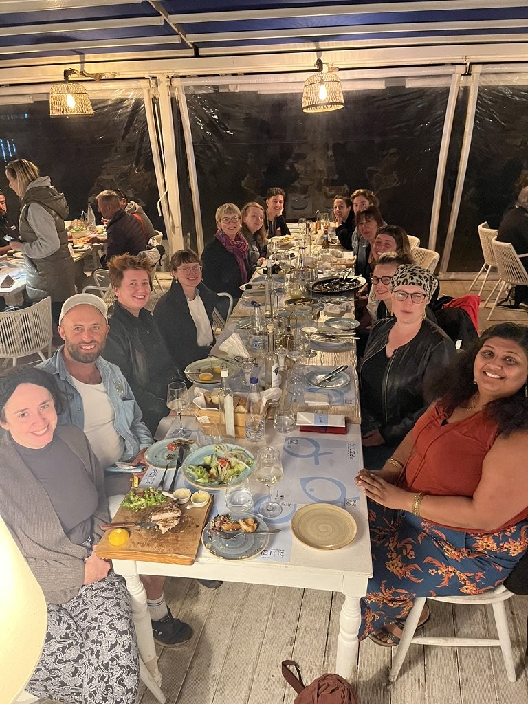 A group of people enjoying a dinner at a long table in a restaurant with warm lighting and large windows.