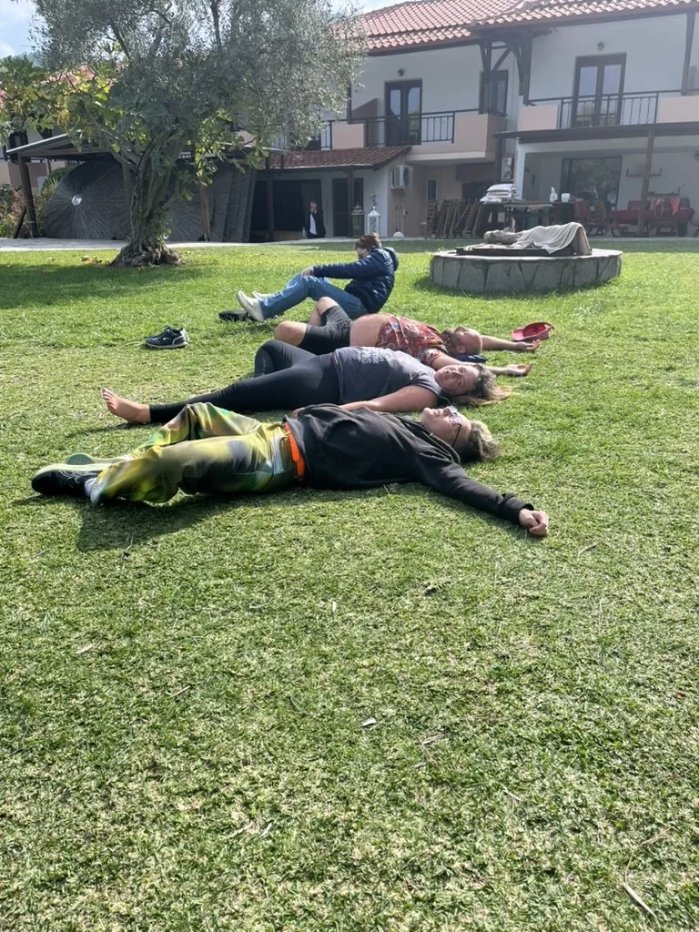 Four people lying on the grass outdoors in a row, with shoes nearby, beside a tree and a round stone structure, in front of a building with balconies.