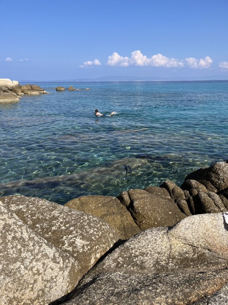 Person swimming in clear ocean water near rocks on the shoreline with a blue sky and clouds in the background.