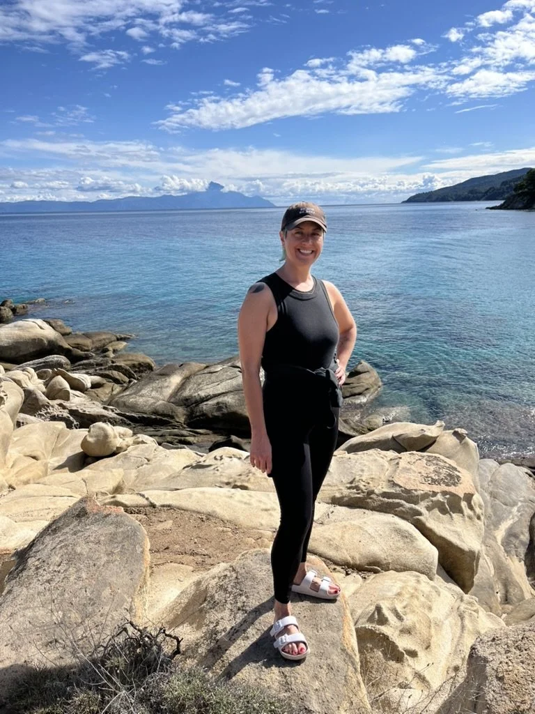 Woman standing on rocks near a body of water with mountains and a partly cloudy sky in the background