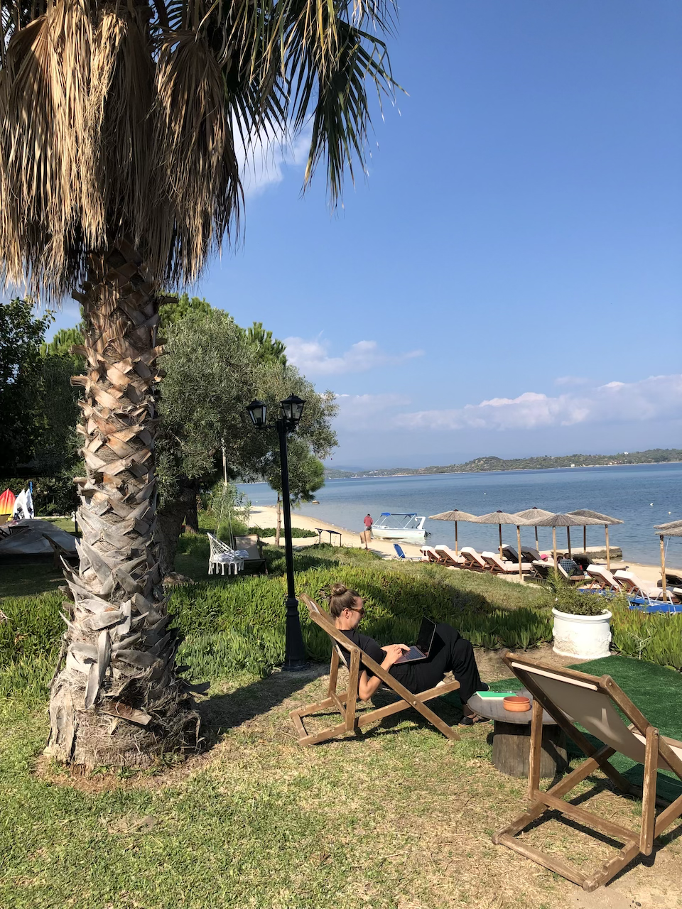 A writer reading and relaxing on a wooden lounge chair under a palm tree, working on a laptop near a beach with umbrellas and lounge chairs, facing the Aegean Sea and a clear blue sky.