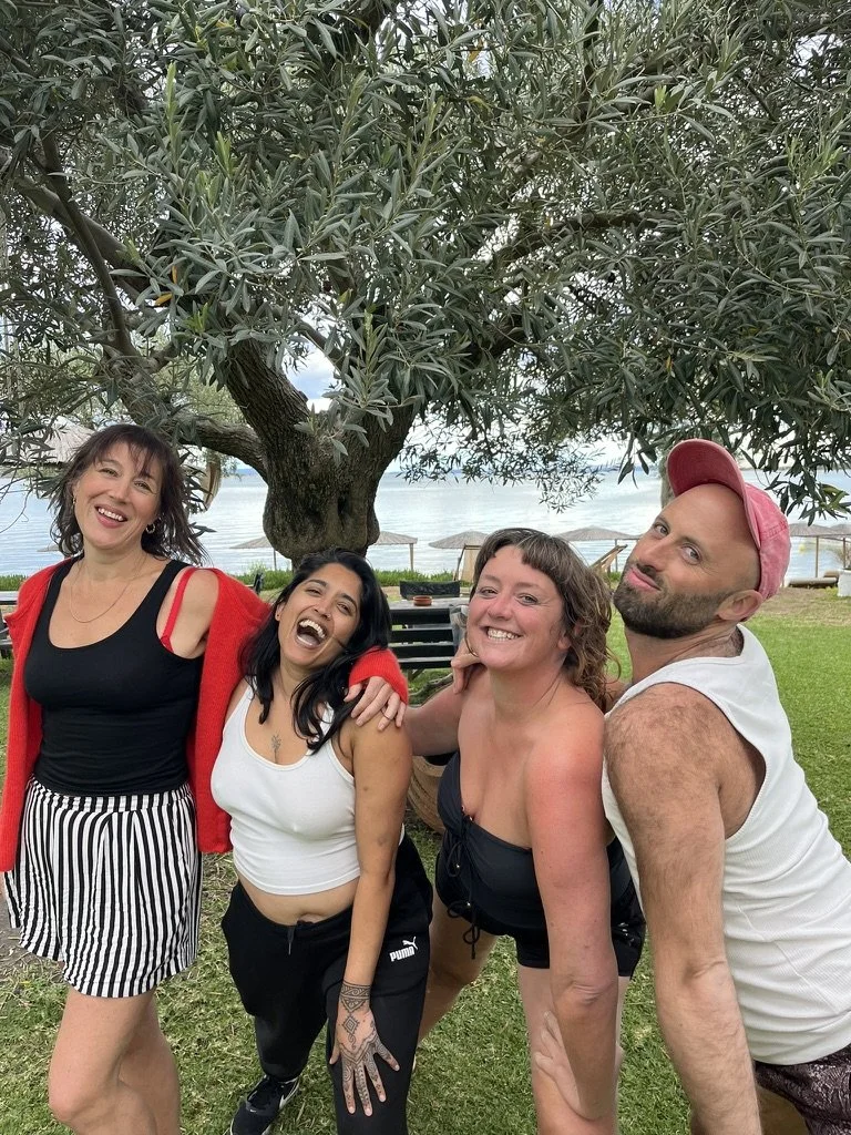 Group of four people smiling and posing outdoors near a lake, with a large tree and umbrellas in the background.