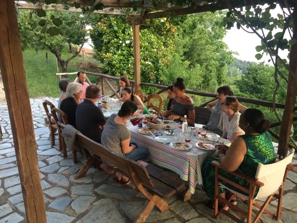 A group of writers sitting around an outdoor dining table on a covered patio, surrounded by green trees and nature at one of Ink and Salt's writing retreats