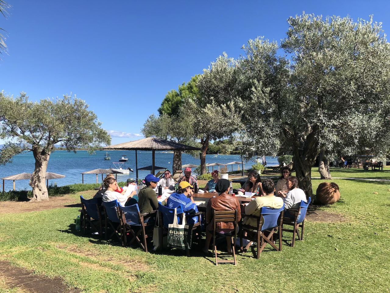 An Ink and Salt writing workshop in progress a large outdoor table by the sea, with trees, umbrellas, and boats in the background on a sunny day. 
