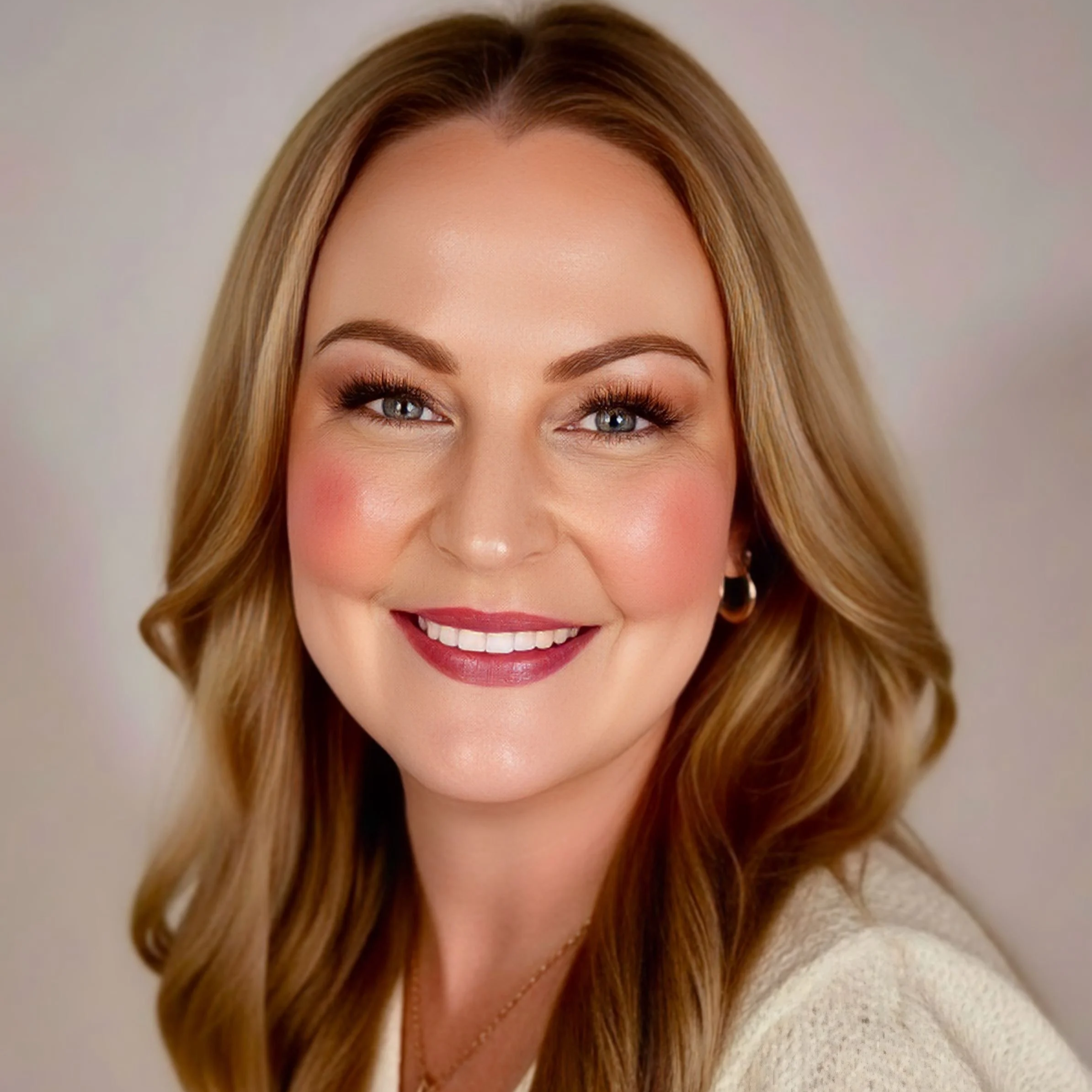 Close-up portrait of a smiling woman with shoulder-length light brown hair, blue eyes, and makeup, wearing a beige sweater and gold jewelry.
