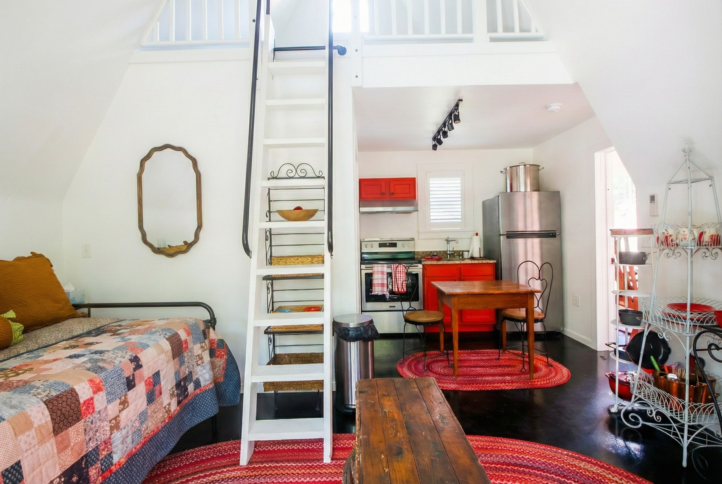 A small living space with a lofted bedroom area, a kitchenette with red cabinets, a stainless steel refrigerator, a wooden dining table with two chairs, a quilted bed with brown pillows, a wooden coffee table, red rugs, and a metal shelving unit with