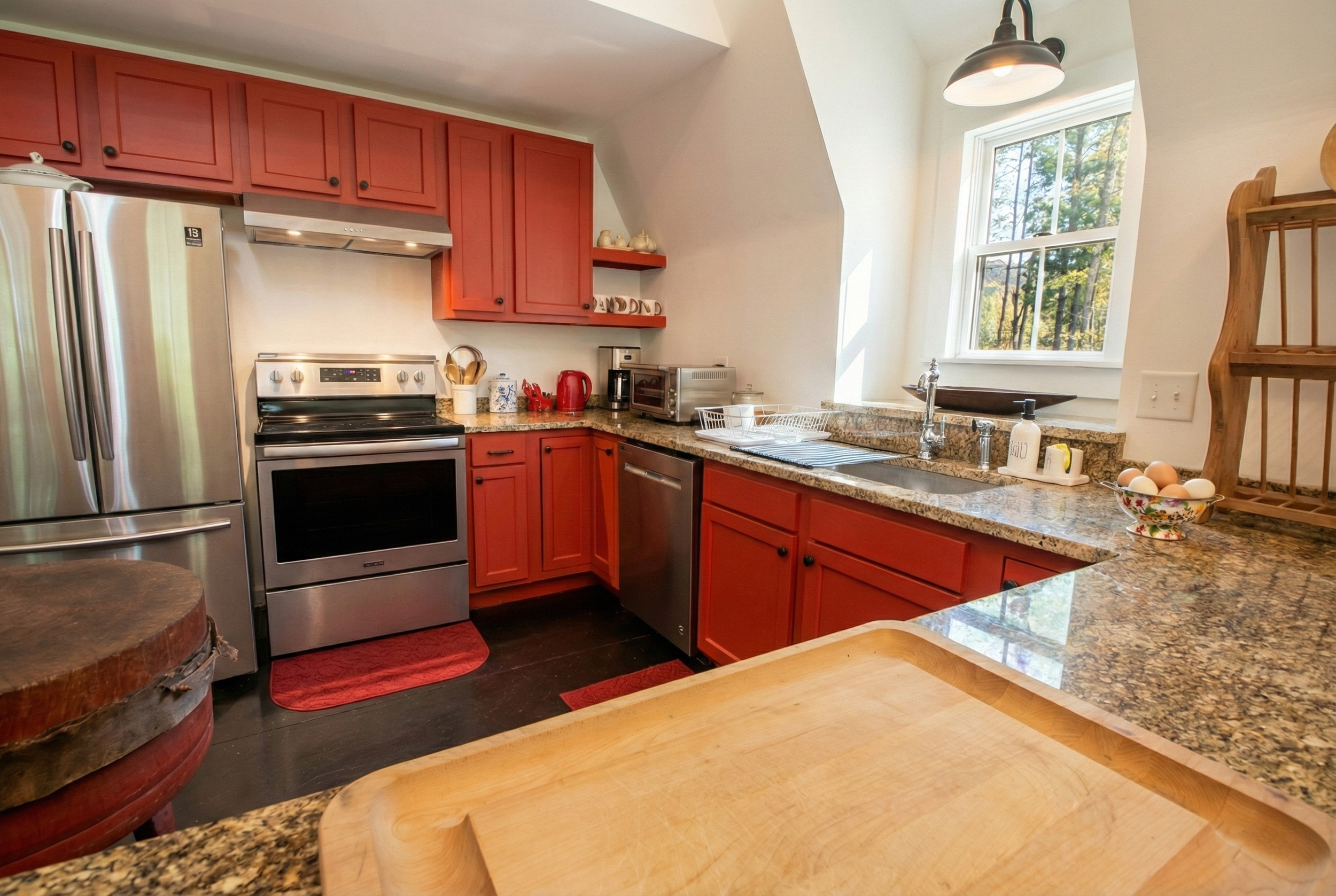 Kitchen with red cabinets, stainless steel refrigerator and oven, granite countertops, window with view of trees, and various kitchen items.