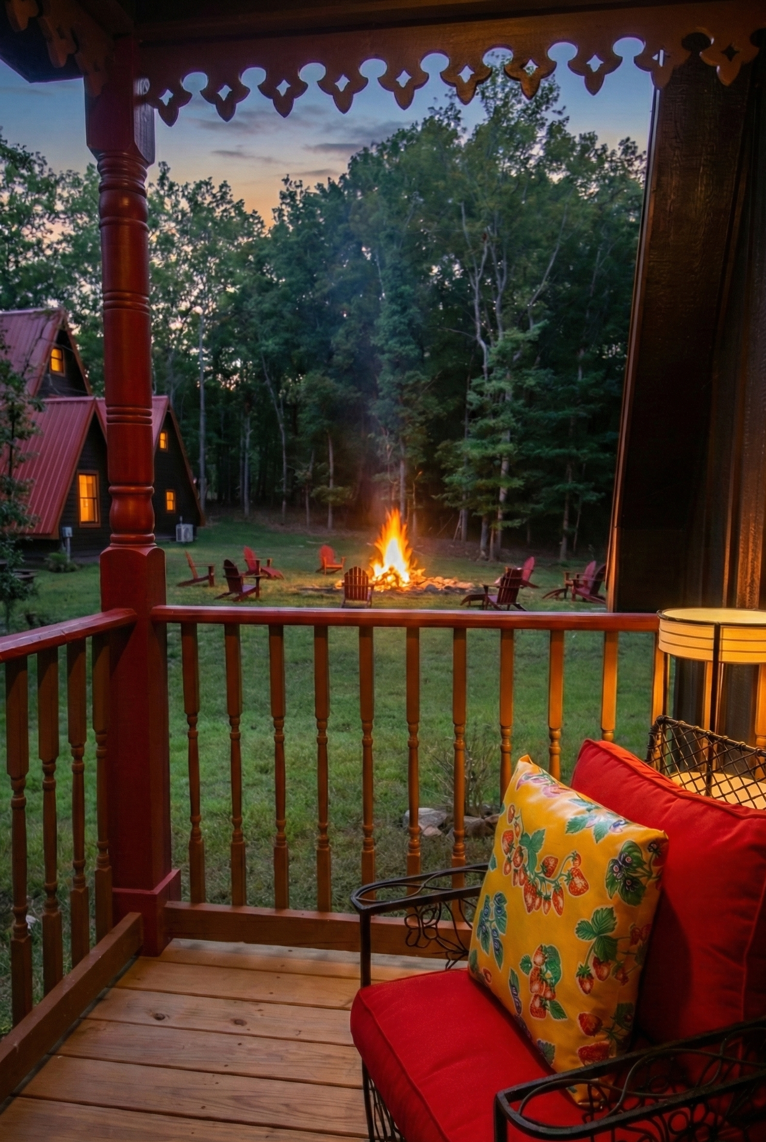 View from a wooden porch looking out at a backyard with a campfire surrounded by Adirondack chairs, trees in the background, and cozy cabin houses with lit windows on a dusk or evening sky.