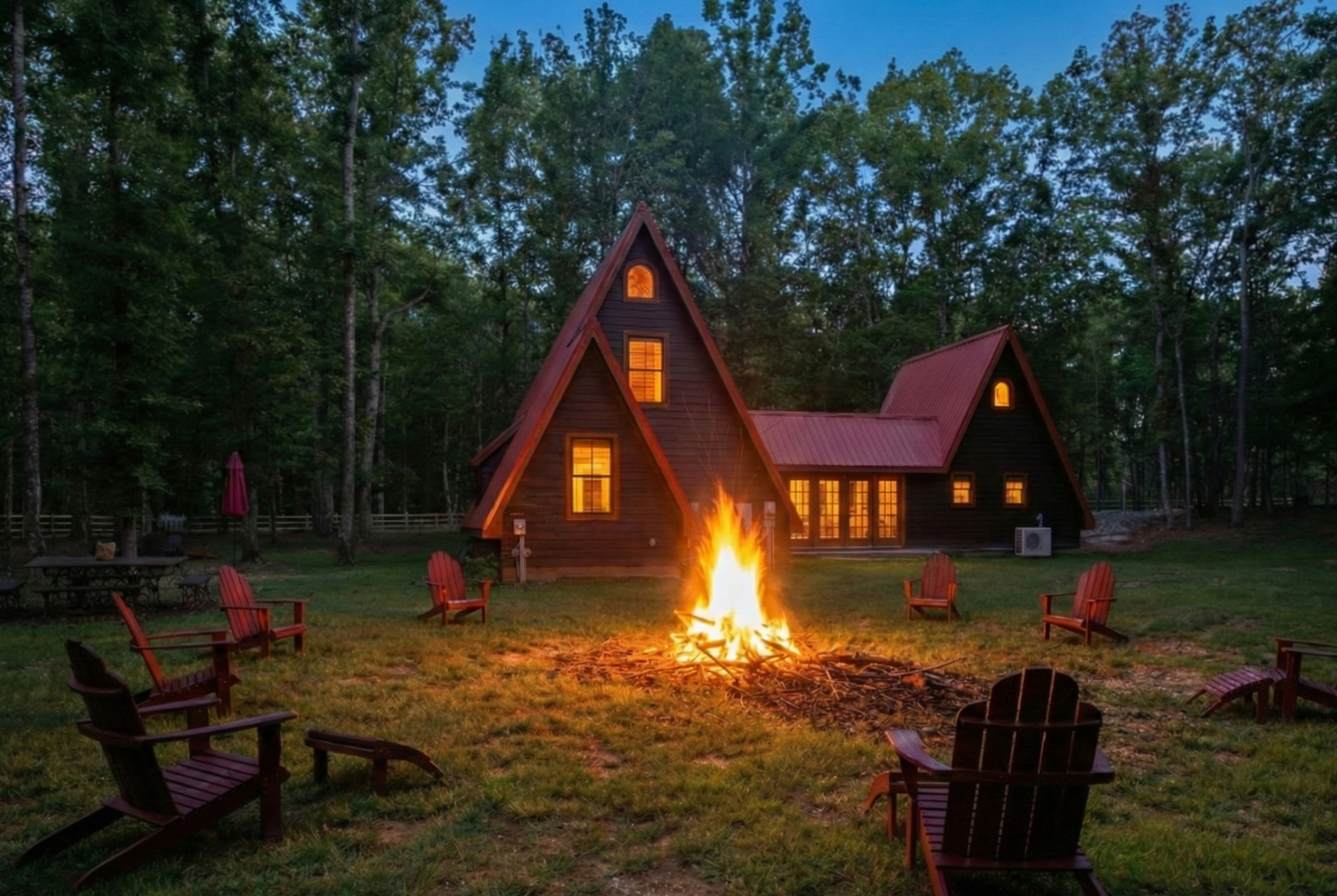 A rustic wooden house with warm lit windows, surrounded by tall trees, with a campfire burning in the yard and red Adirondack chairs arranged around it at dusk.