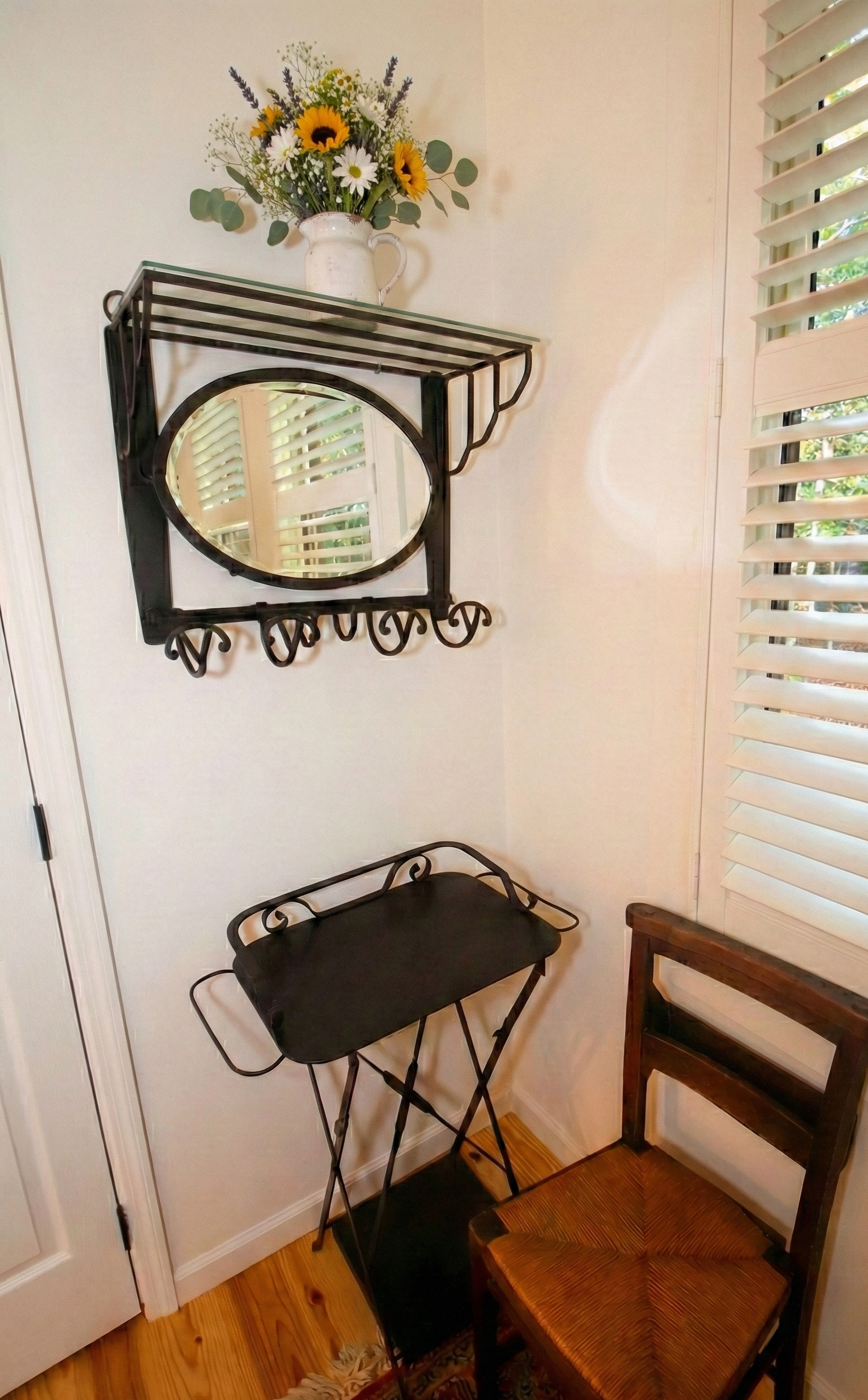 A small corner with a wall-mounted black metal shelf holding a white pitcher vase filled with sunflowers, daisies, and other flowers, a black foldable tray table, a wooden chair with a woven seat, and windows with white plantation shutters.
