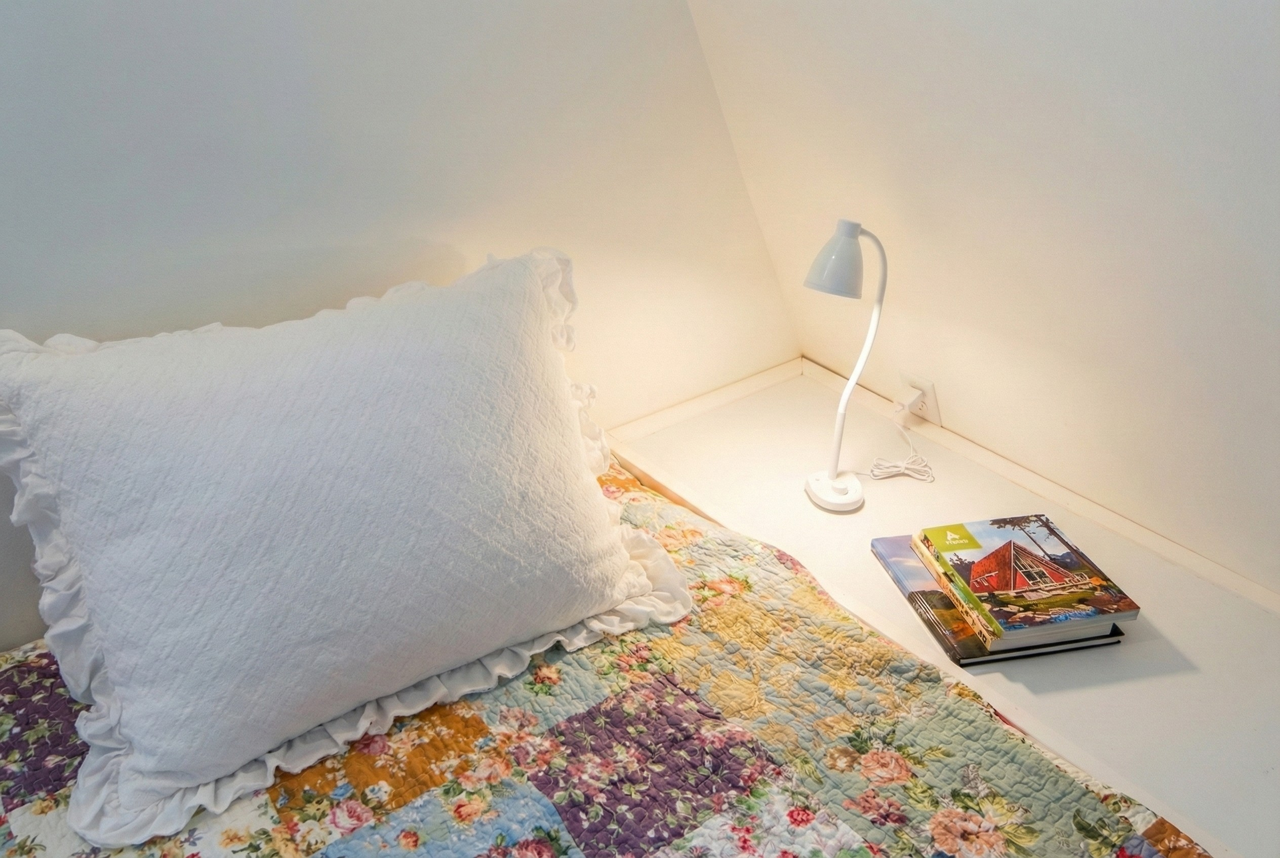 A bedside table with a white table lamp and three books, next to a bed with a floral quilt and a large white pillow.