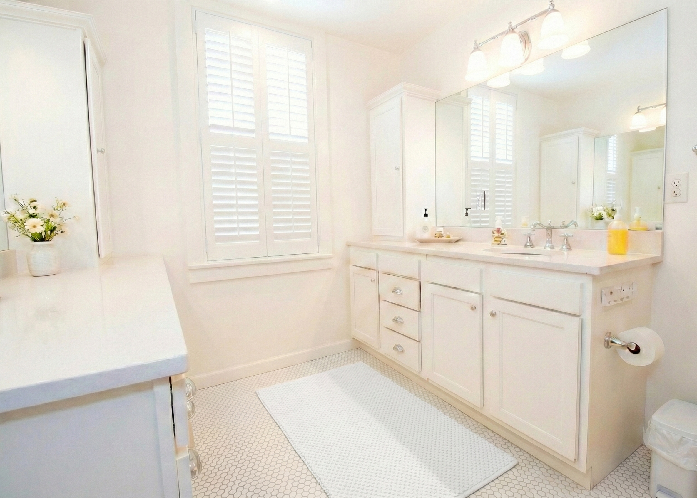 Bright white bathroom with double sink vanity, large mirror, plantation shutters on window, white cabinets, and a white rug on white hexagonal tiled floor.