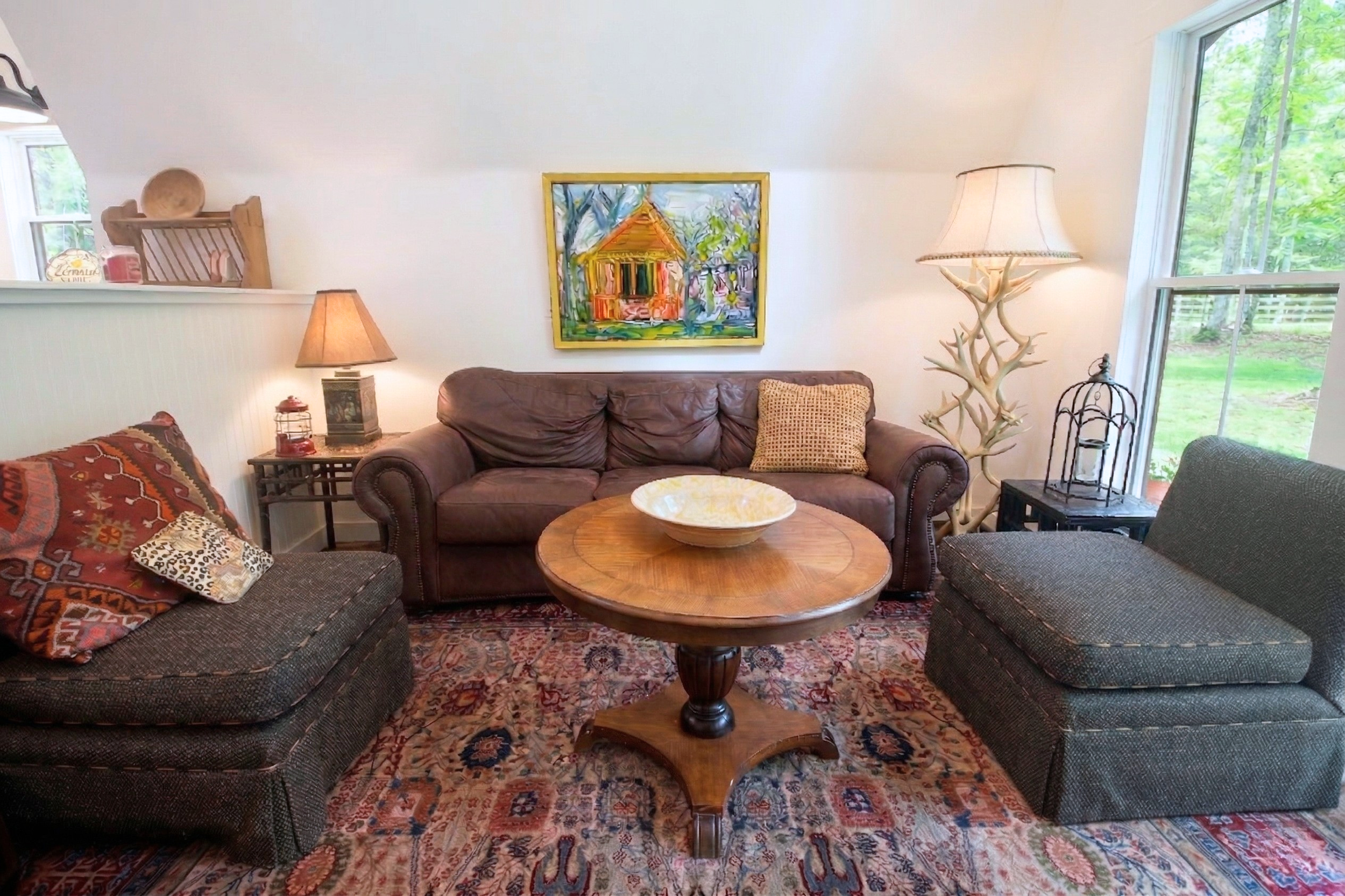 Cozy living room with a brown leather sofa, two patterned armchairs, a round wooden coffee table, colorful area rug, and various lamps and decorations, with a window showing green outdoor scenery.