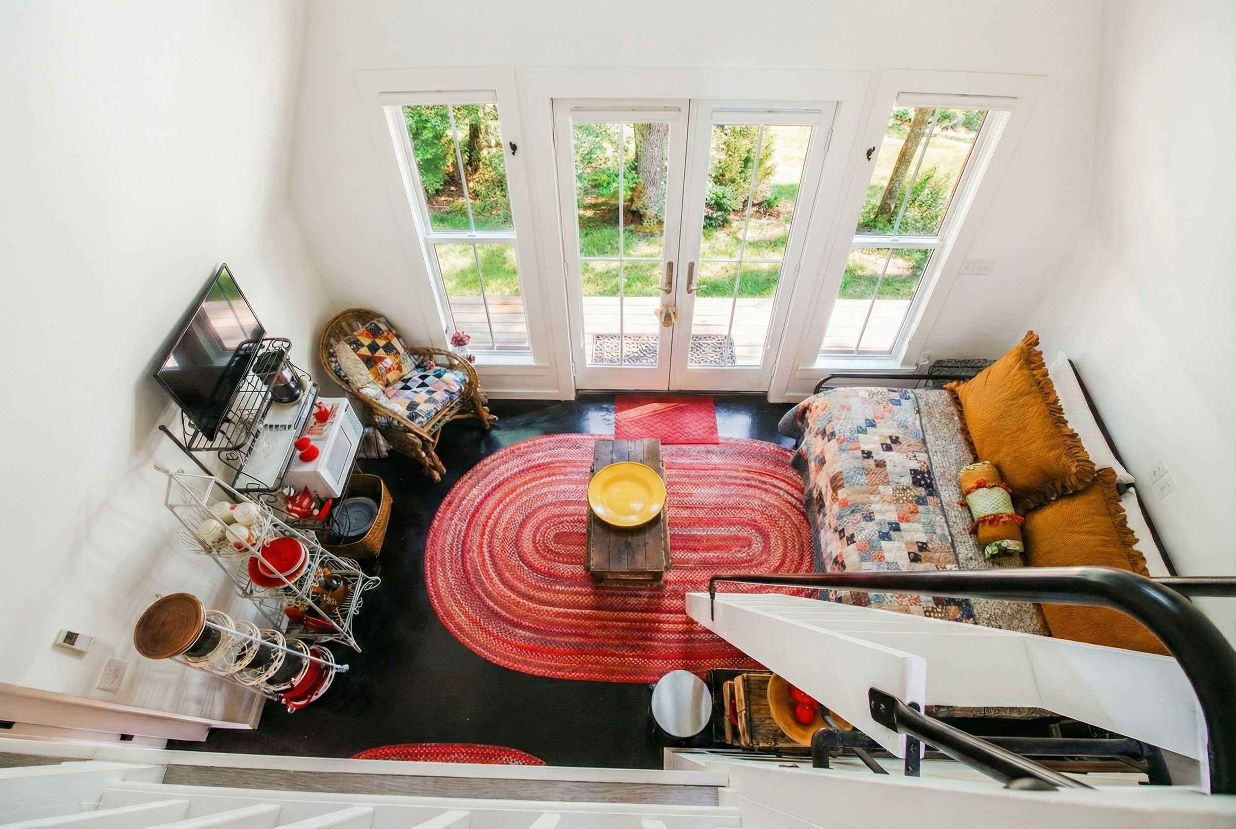 Living room with large glass doors leading to a green outdoor area, featuring a patterned sofa with yellow cushions, a round red rug, a wooden coffee table with a yellow decorative bowl, a wicker armchair with a quilted cushion, a small white cart wi