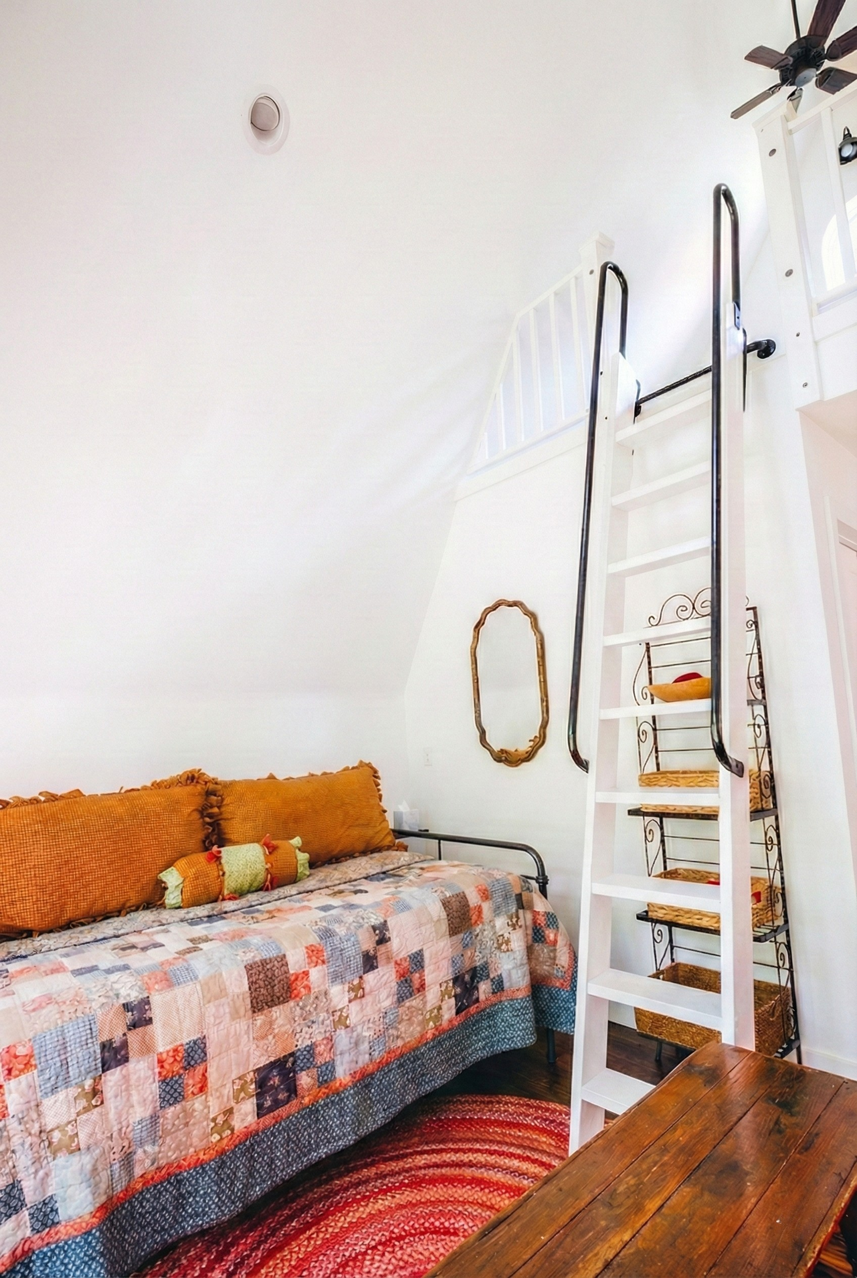 A cozy room with a quilt-covered bed, orange pillows, a colorful striped pillow, a large wooden table, a decorative mirror, and a white ladder leading to a loft area with black safety rails. The room has white walls and a ceiling fan.