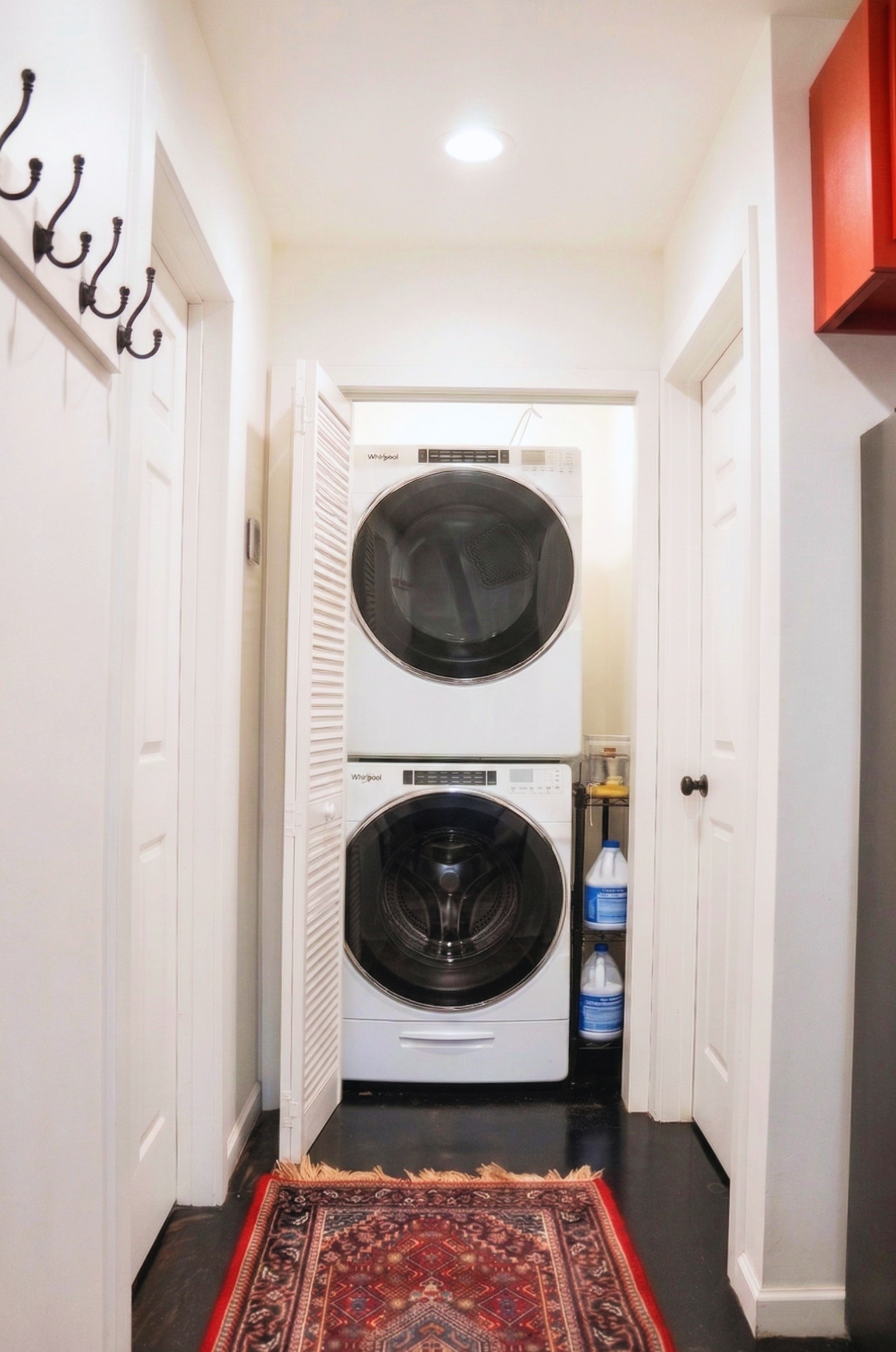 Stacked white washer and dryer in a small laundry area with cleaning supplies stored to the side, a red patterned rug on a dark floor, and white walls with doors and hooks.