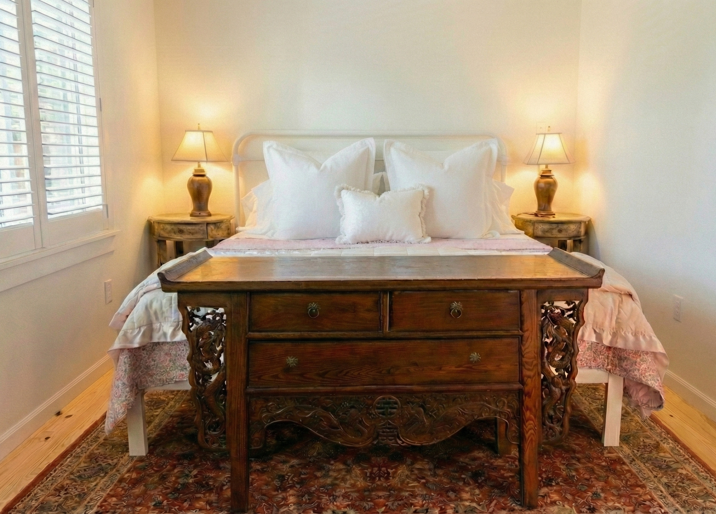 A cozy bedroom with a bed, decorative pillows, nightstands with lamps, a wooden bench at the foot of the bed, and a rug on the floor.