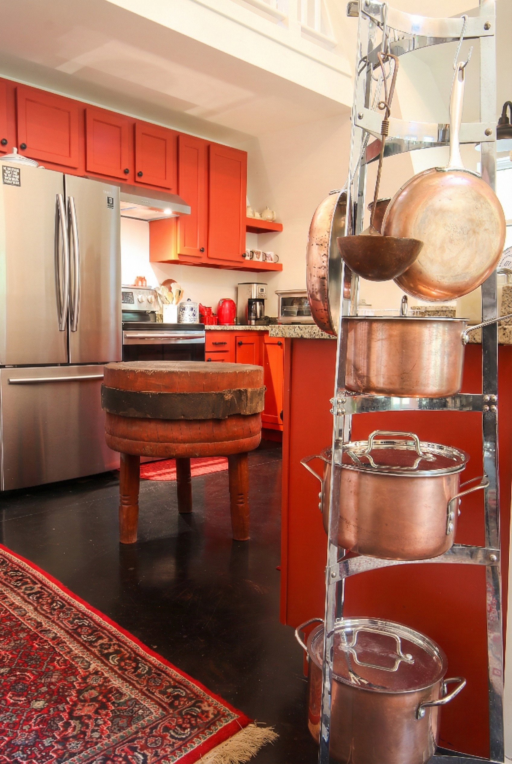 Kitchen with red cabinets, stainless steel refrigerator, and a metal rack holding copper pots and pans. An old wooden stool, a patterned rug, and various kitchen appliances are visible.