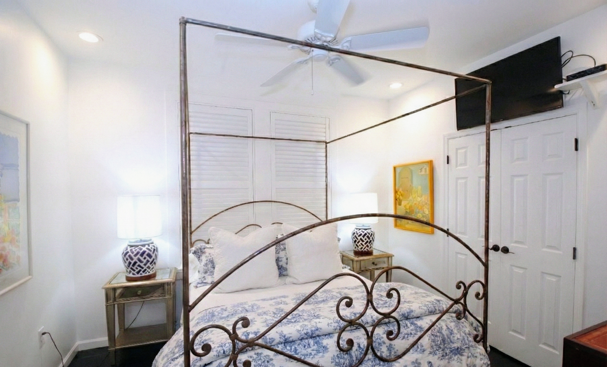 A bedroom with a wrought iron bed, white bedding with blue floral pattern, nightstands with matching lamps, white walls, a painting, a television mounted above the door, and closed white closet doors.