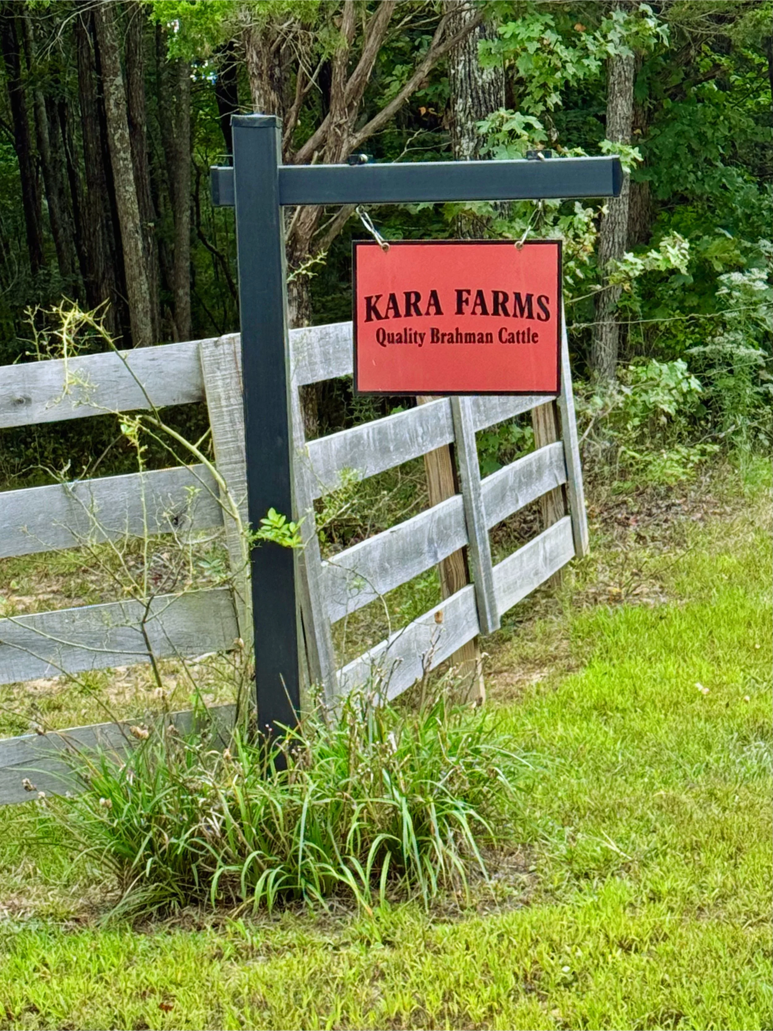 Sign for Kara Farms with red background and black text, next to a wooden fence and trees in a grassy yard.