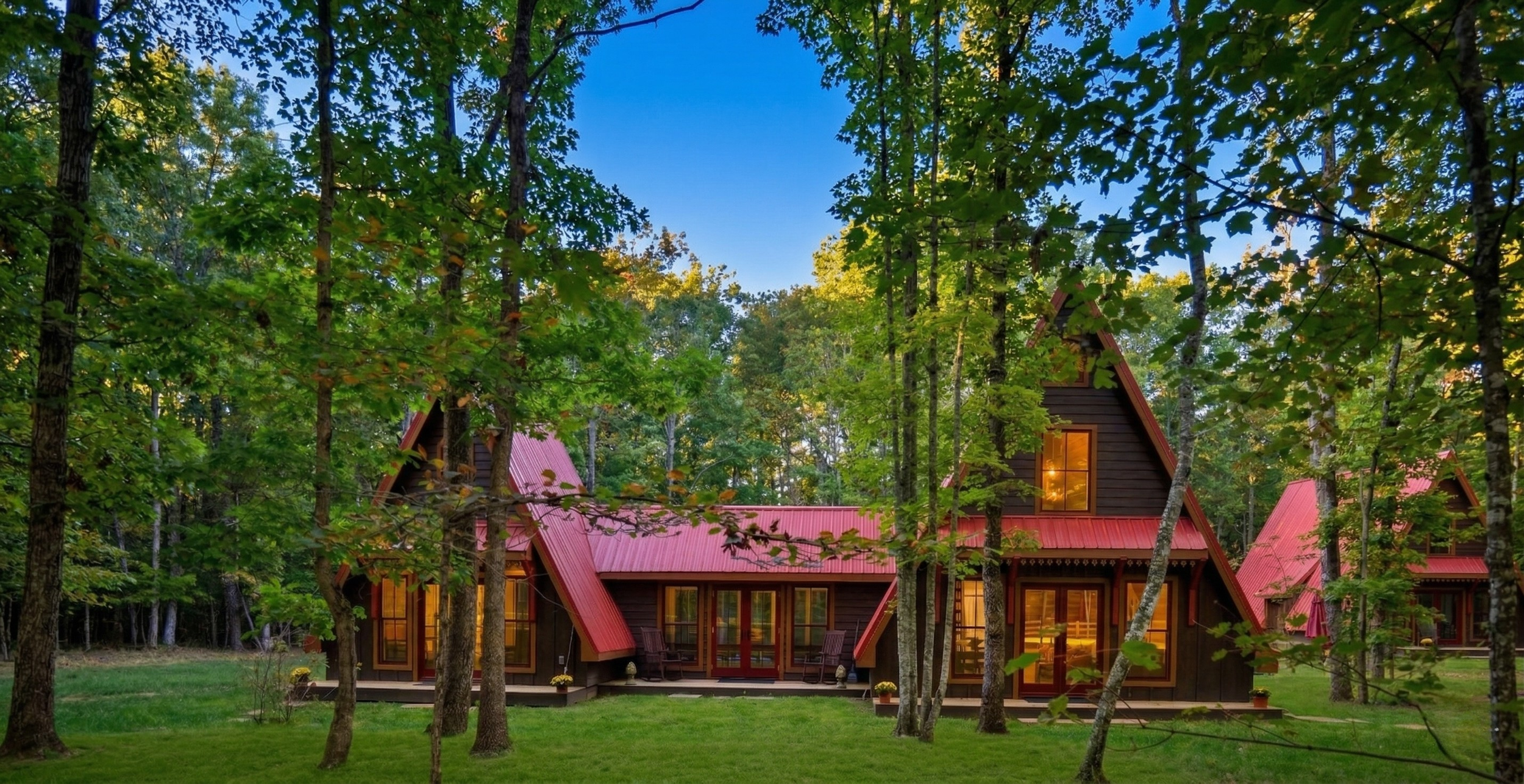 A cabin with a red roof in a wooded area with tall trees and green grass, with windows glowing from interior lighting, during twilight.