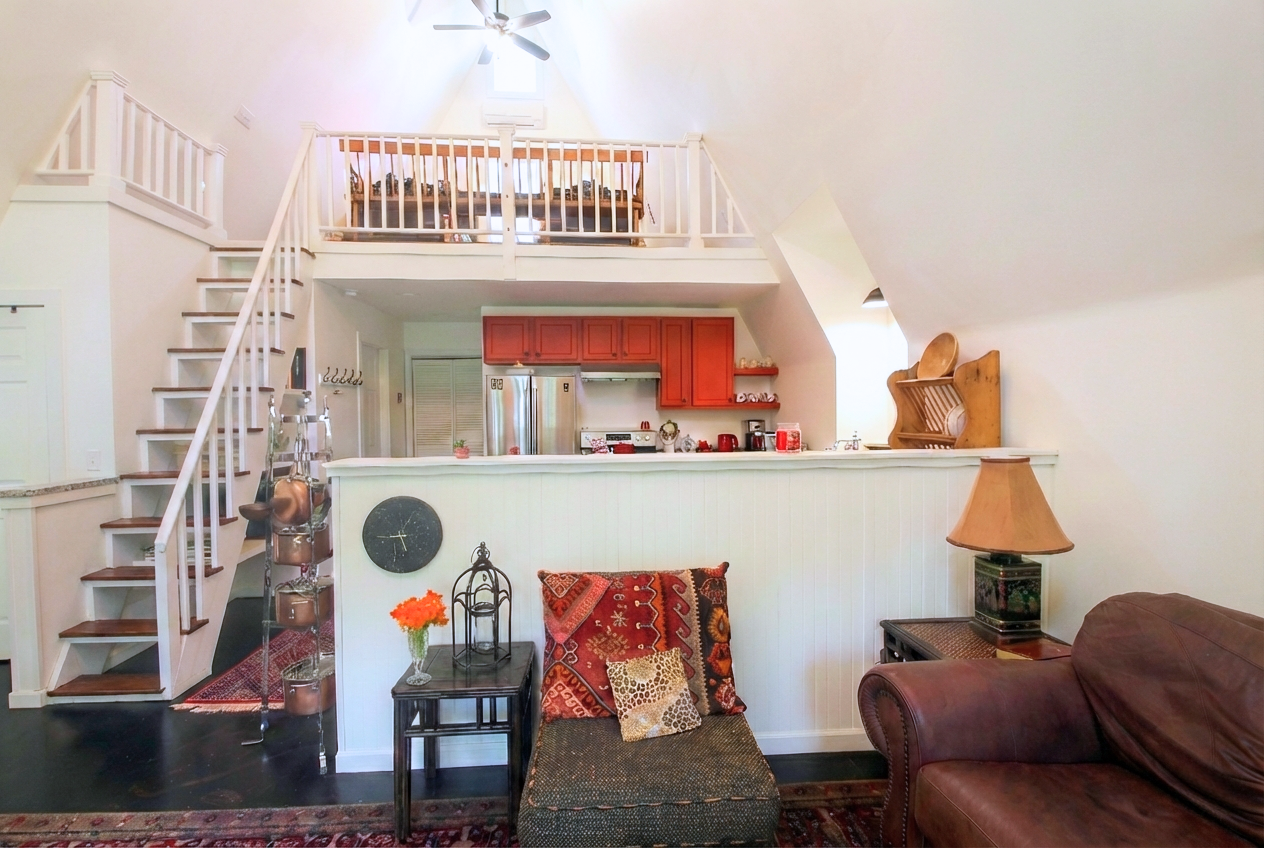 Living room with a staircase leading up to an open balcony. The kitchen with red cabinets is visible behind a half wall. The room has a brown armchair, a small table with a lamp, a black side table with flowers, and decorative pillows. The walls are 