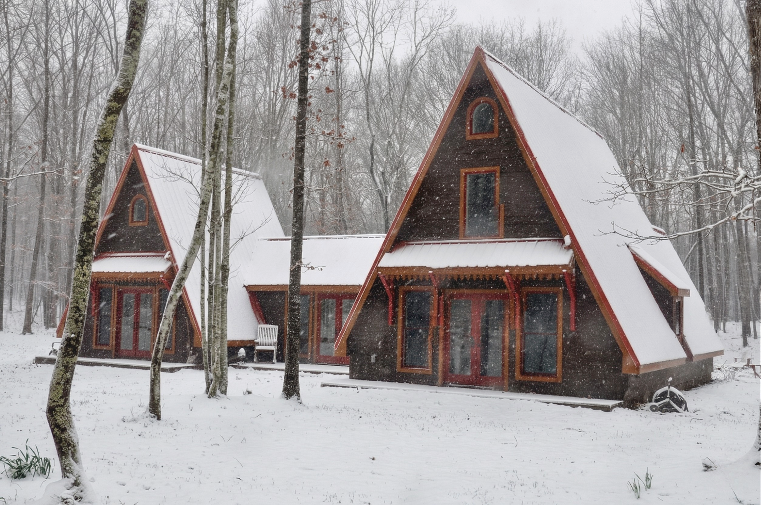 Two A-frame cabins with snow-covered roofs in a snowy, wooded landscape during snowfall.