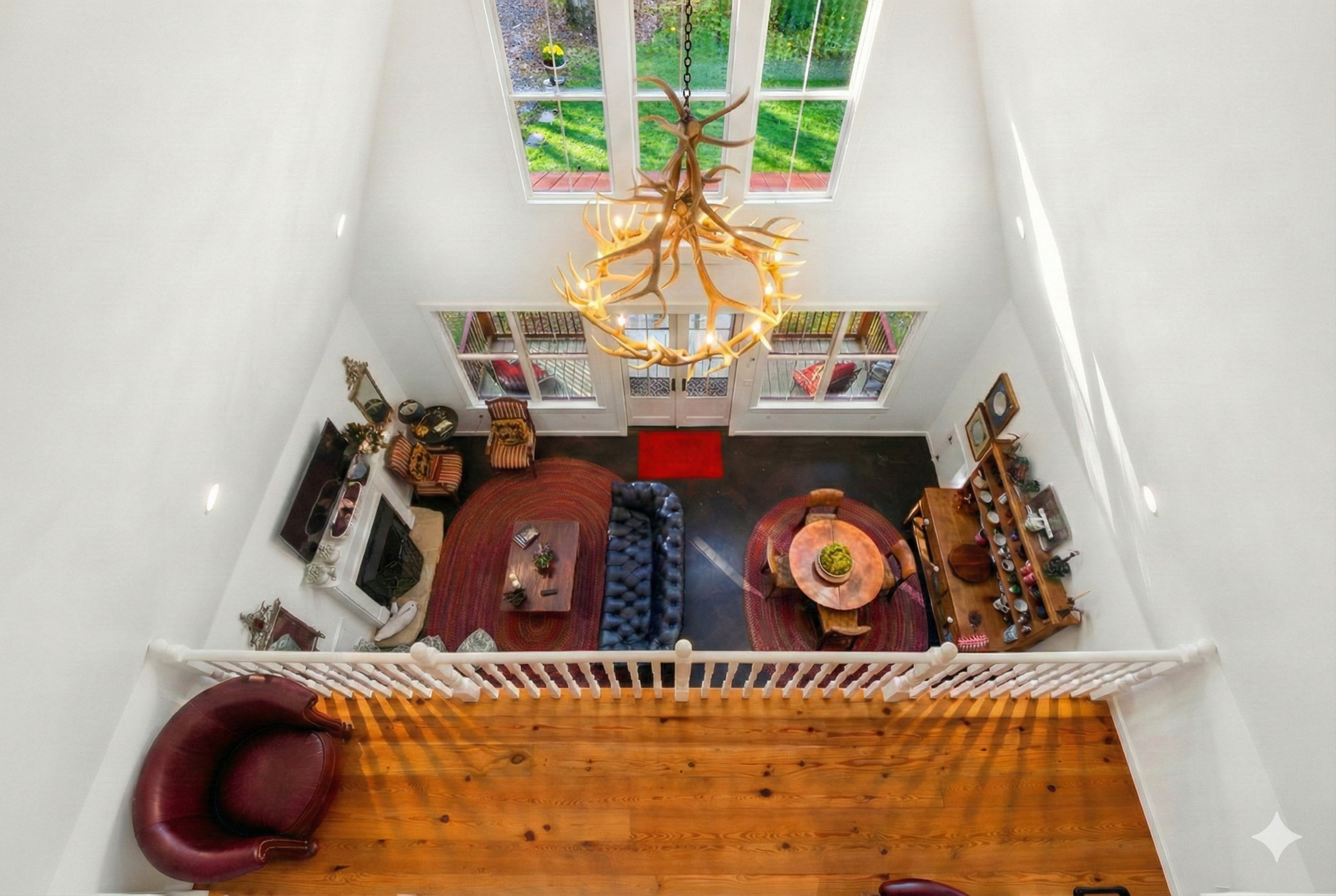 A spacious living room with high ceilings and large windows, featuring a chandelier made of antlers hanging from the ceiling, a red rug, a sofa, a round dining table with chairs, a TV, and wooden furniture, with a balcony visible through the windows.